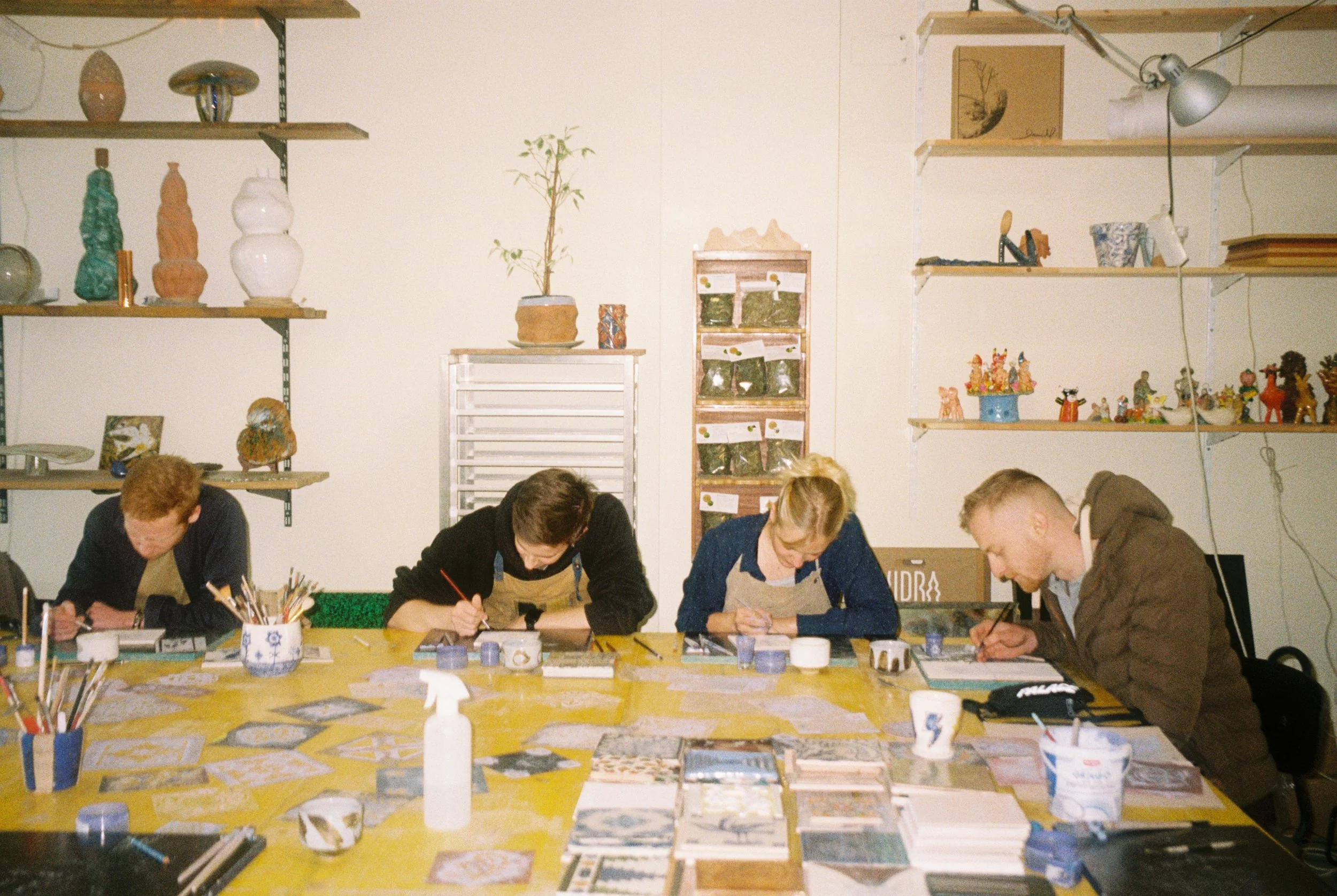 Vier mensen zitten aan een grote tafel die bedekt is met kunstmaterialen, en werken aan schilderijen of tekeningen. Op de tafel staan penselen, potten, en papier. Op de achtergrond bevinden zich naar links en rechts planken met keramiek en knutselobj