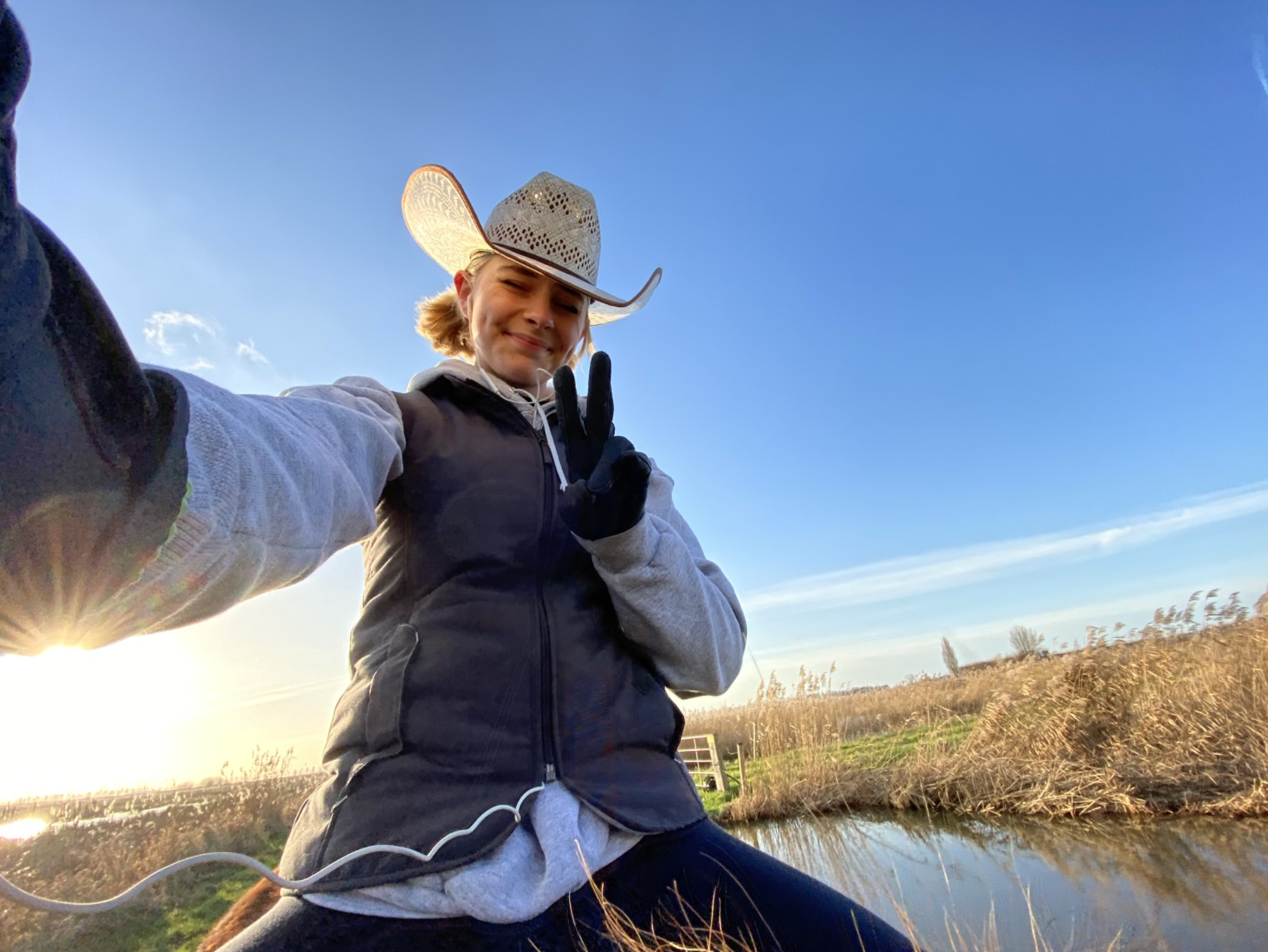 Een vrouw maakt een selfie in een landelijke omgeving met een rivier en akkers, draagt een cowboyhoed, handschoenen, en glimlacht terwijl ze een vredesteken maakt met haar vingers, tijdens zonsondergang of -opkomst.