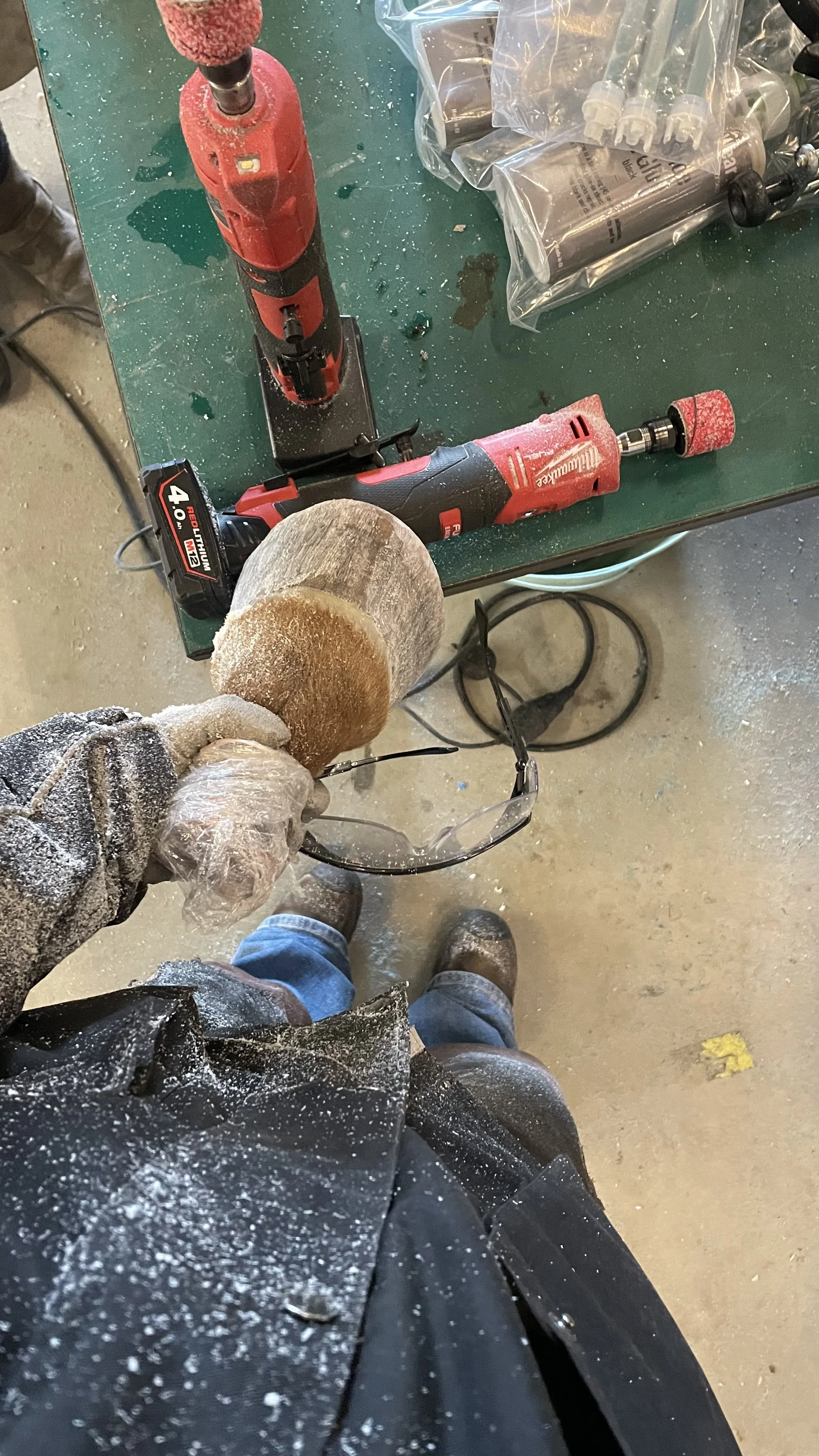 Een persoon houdt een afvalborstel vast boven een werkbank met elektrische gereedschappen, waaronder een rode Milwaukee accuboormachine en een black and decker acculader, omgeven door stof en kleine deeltjes.