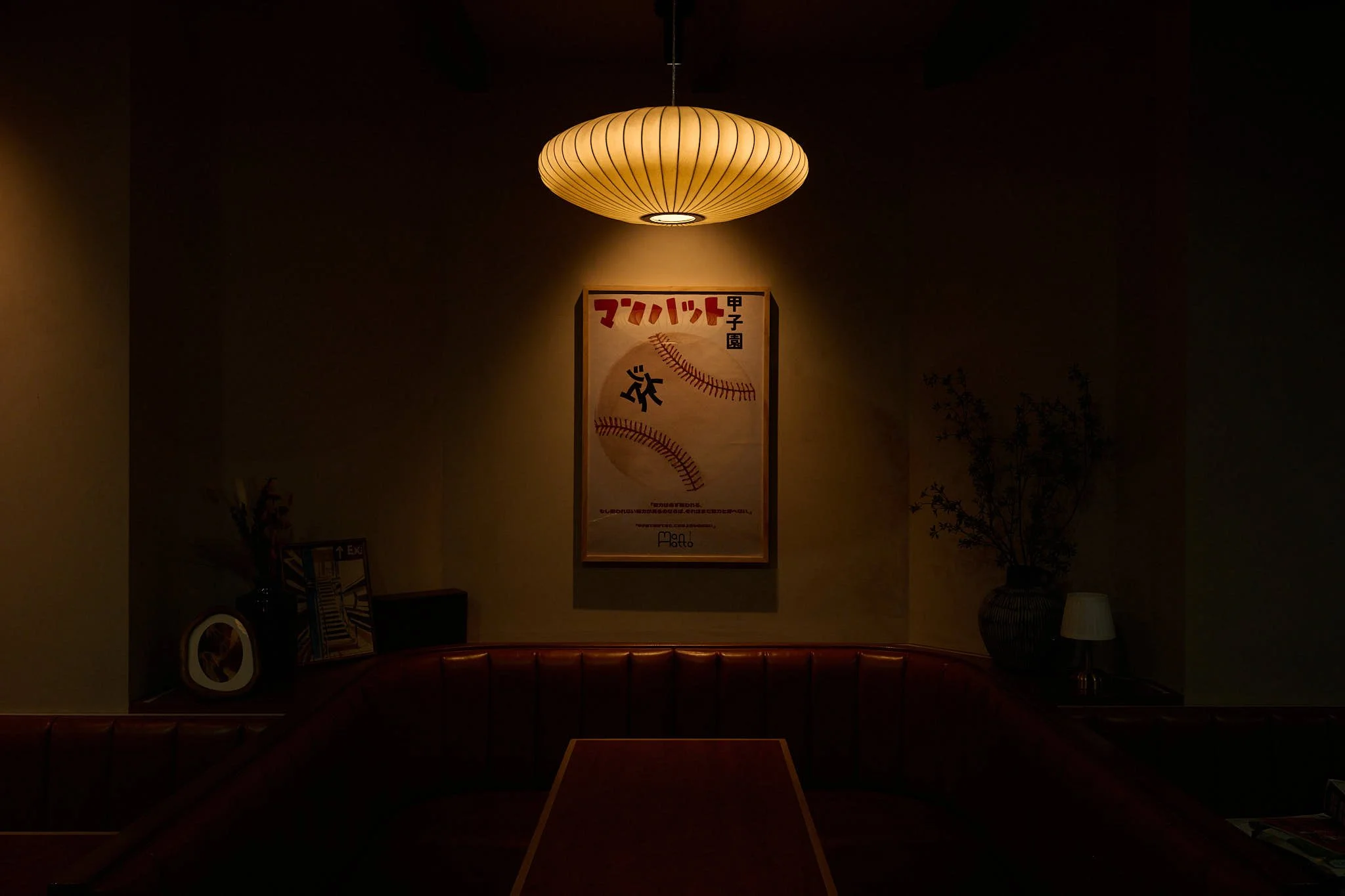 Dimly lit room with a round ceiling lamp and framed Japanese poster on the wall, with a corner shelf holding decor items and a large vase with branches.