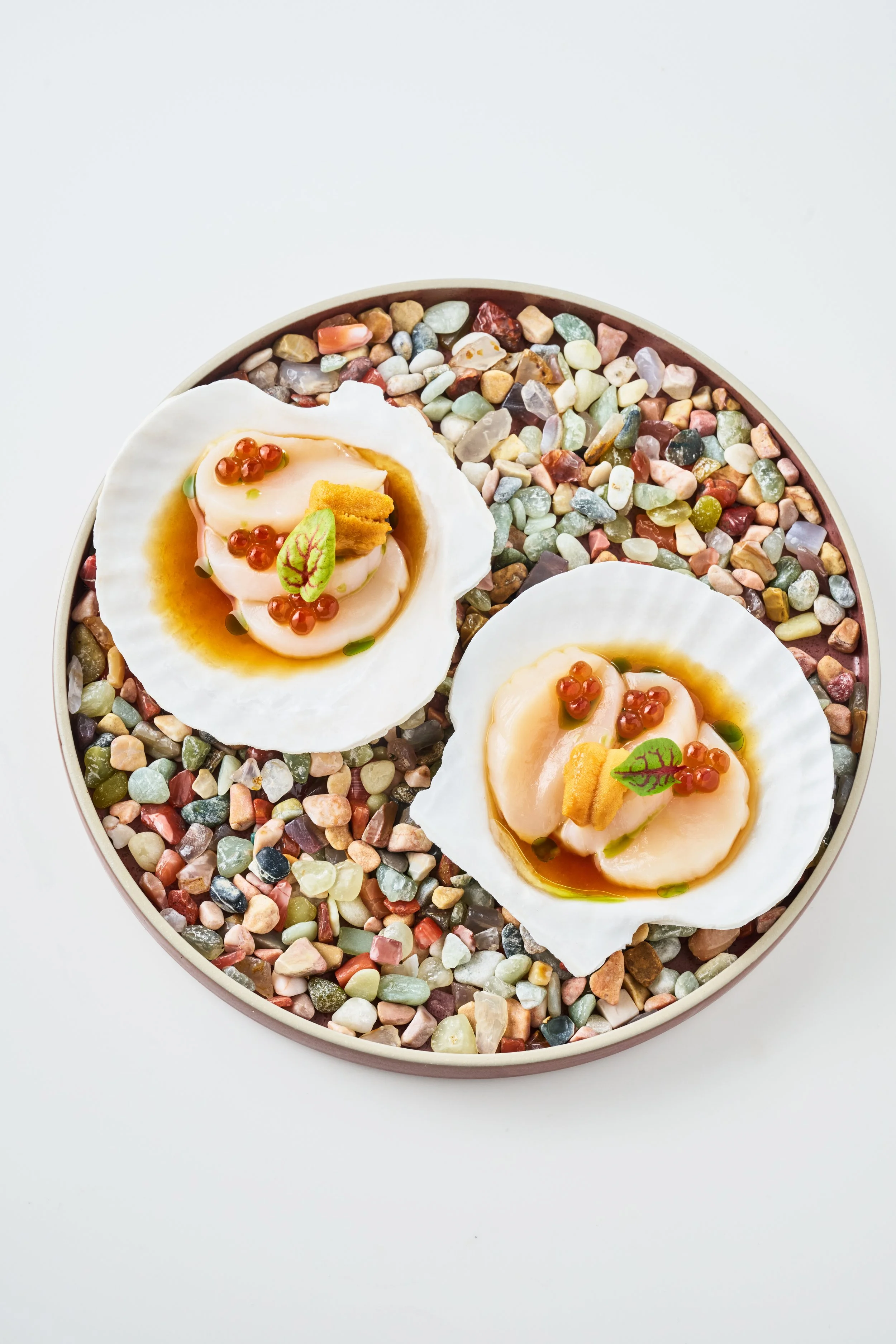 Two scallops served on shells on a bed of colorful pebbles in a round container.