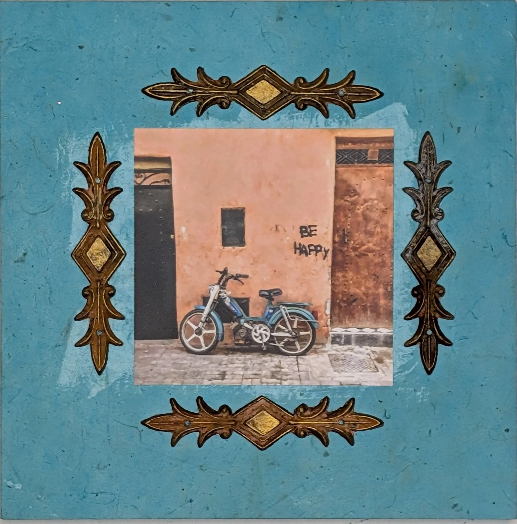 Be Happy is an 8x8 collage on board that merges photography with vintage flair and textured embellishment. Captured in a quiet alley with sun-washed coral walls and a resting moped, the scene is anchored by the graffiti message: “Be Happy.” Framed wi