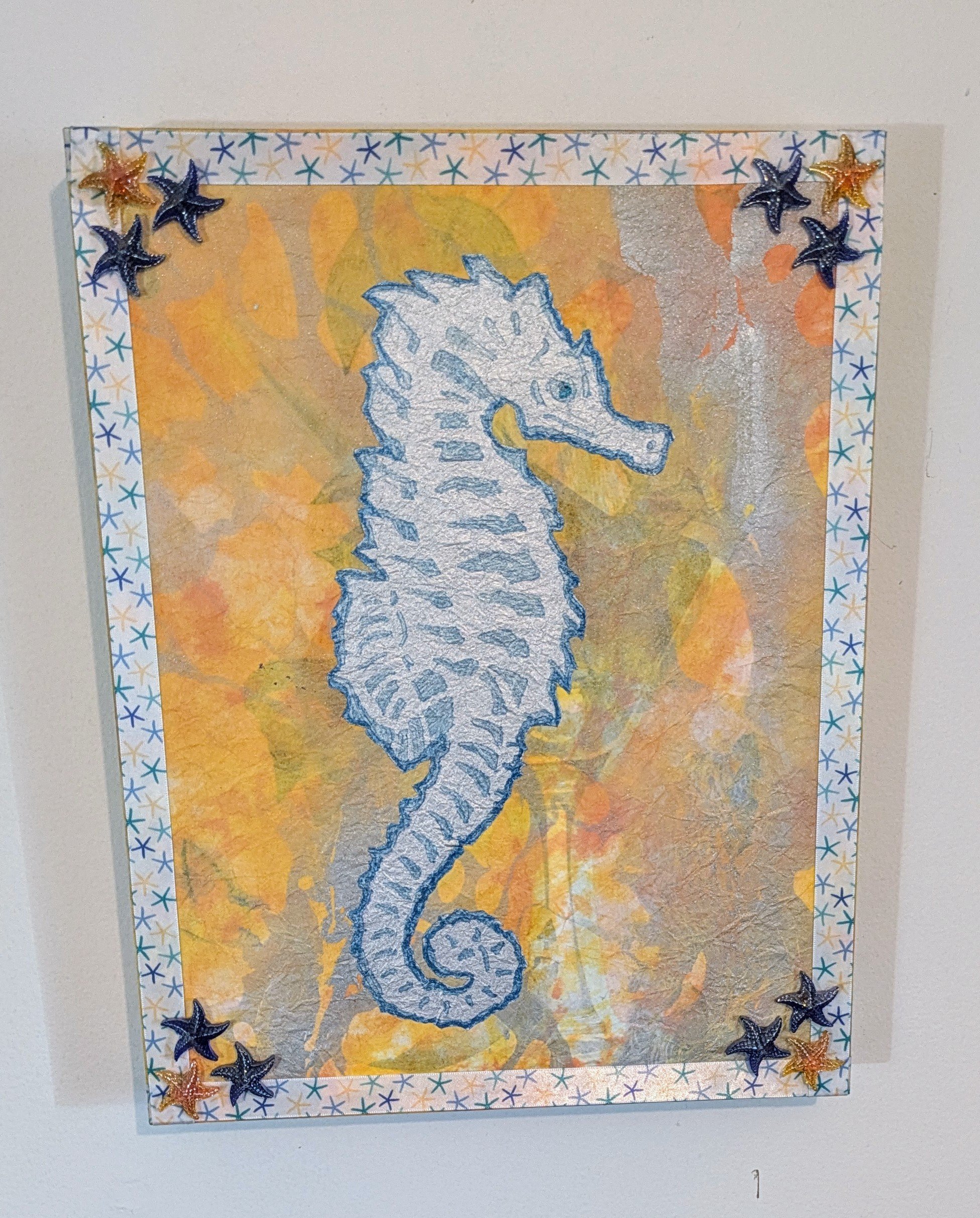 Silver Seahorse is an original mixed media collage on board, featuring a shimmering seahorse silhouette set against a warm, abstract ocean background. Embellished with metallic accents, layered textures, and decorative starfish corners, this piece ev