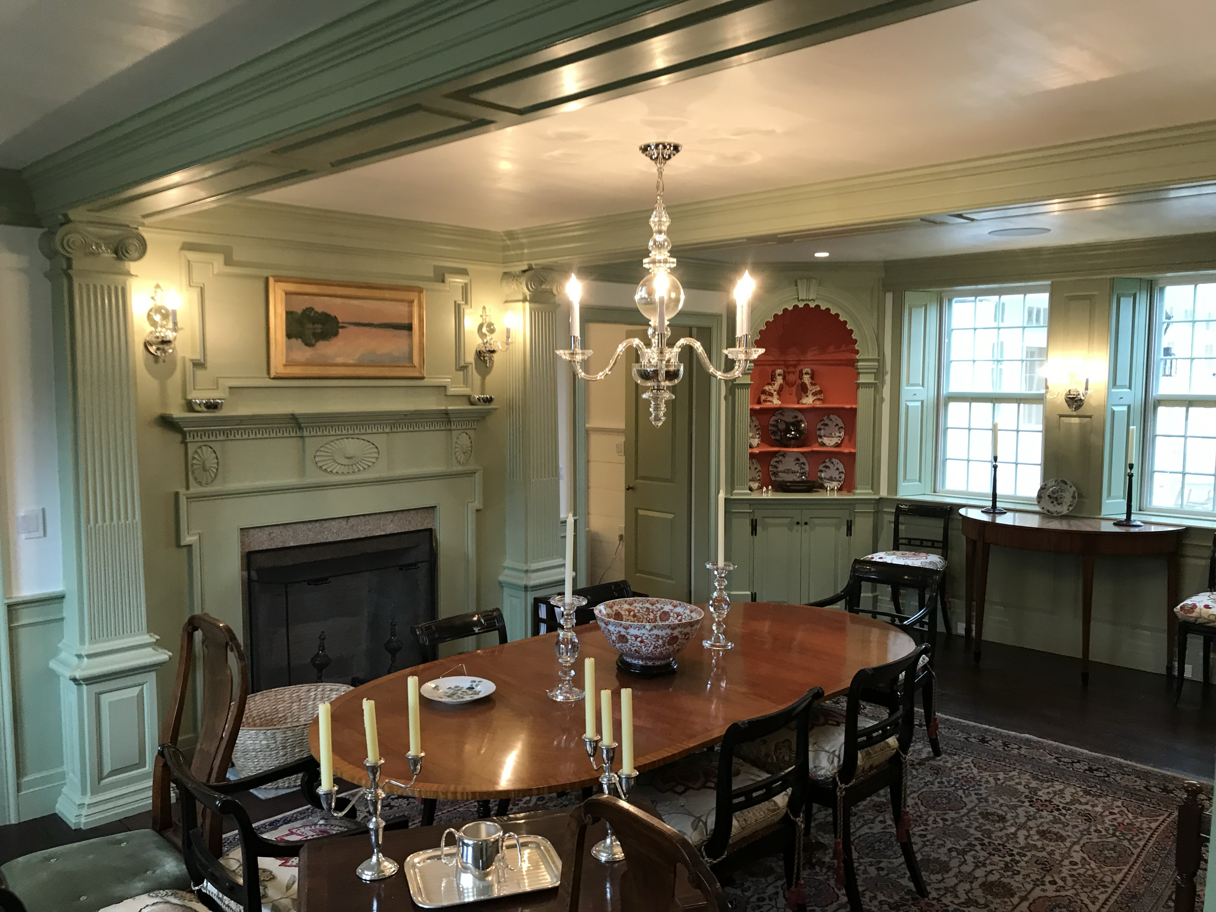 A dining room with a large wooden table set with candles and a decorative bowl, surrounded by chairs, with a chandelier hanging above. The room has a fireplace, built-in shelves with plates, a window allowing natural light, and is decorated with pain