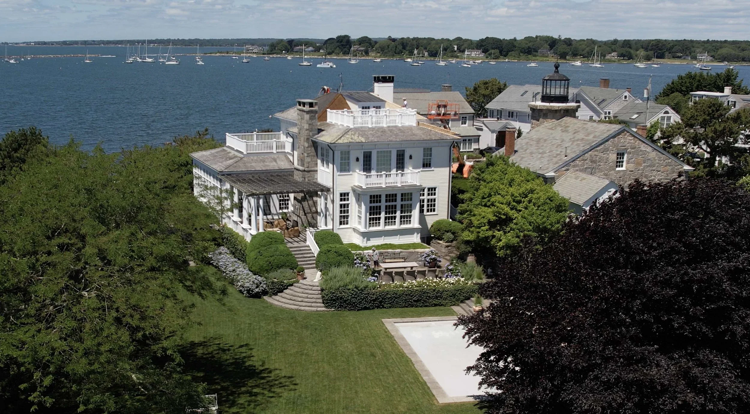 Aerial view of a waterfront residential area with multiple houses, lush greenery, a backyard with a terrace, and a nearby harbor with sailboats.