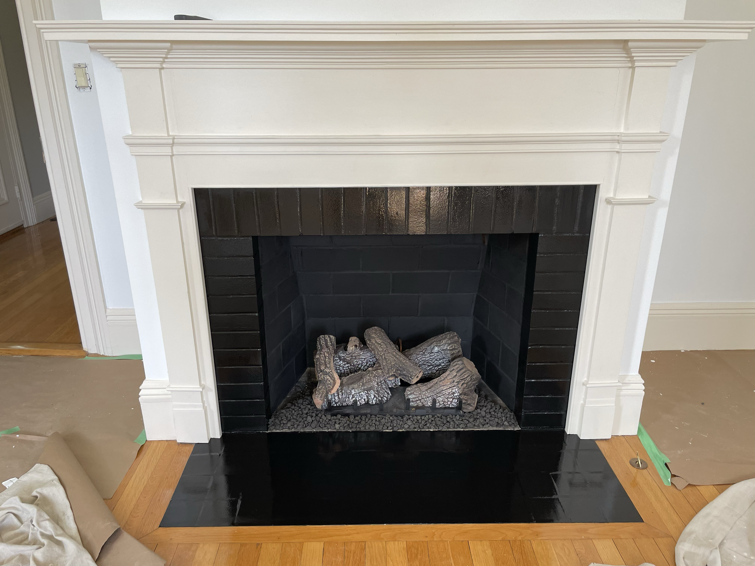 Unfinished fireplace with a white mantel and black interior, containing faux logs and a black surround at the base, surrounded by hardwood floors and some items of packaging and paper on the floor.