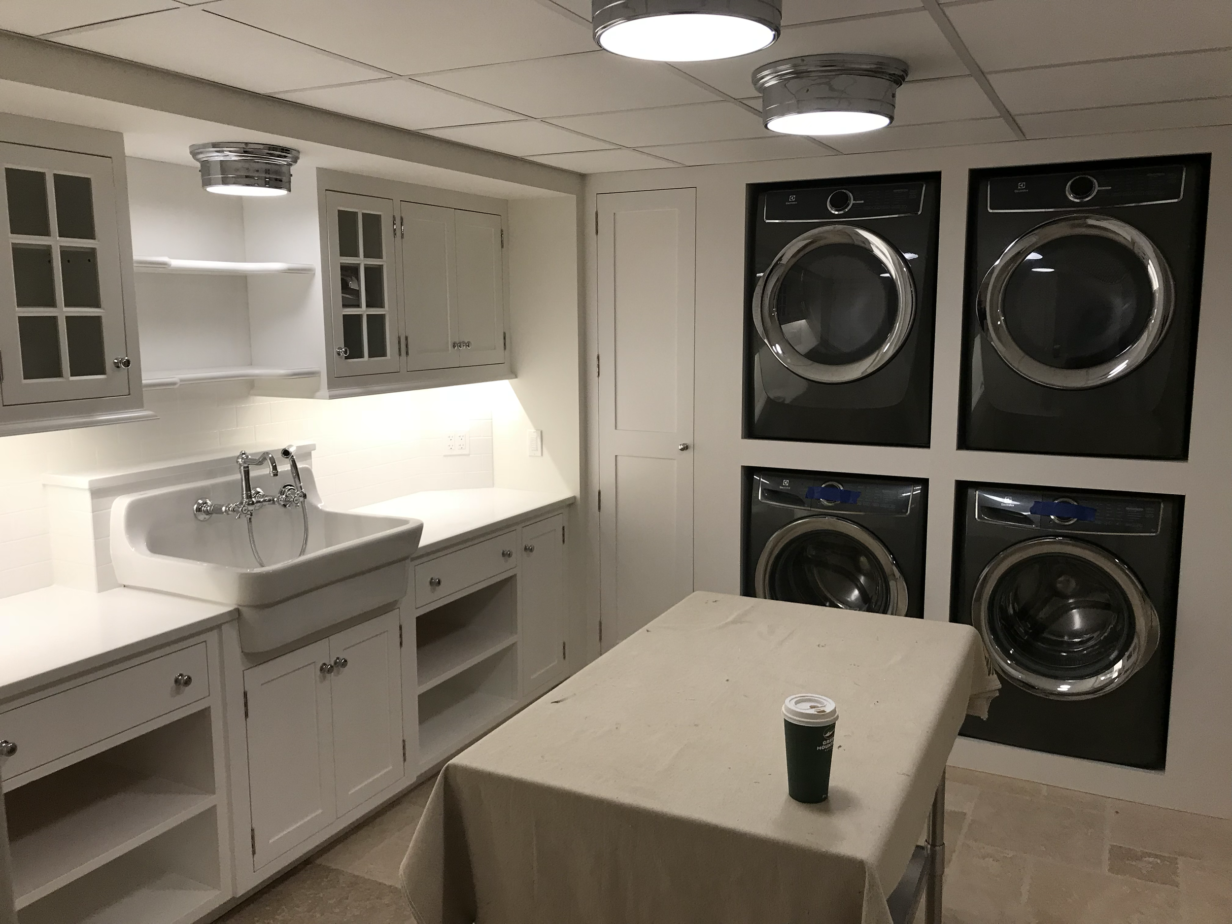 A laundry room with gray cabinets, a white farmhouse sink, a countertop, a small table with a coffee cup, and stacked black front-loading washing machines and dryers.