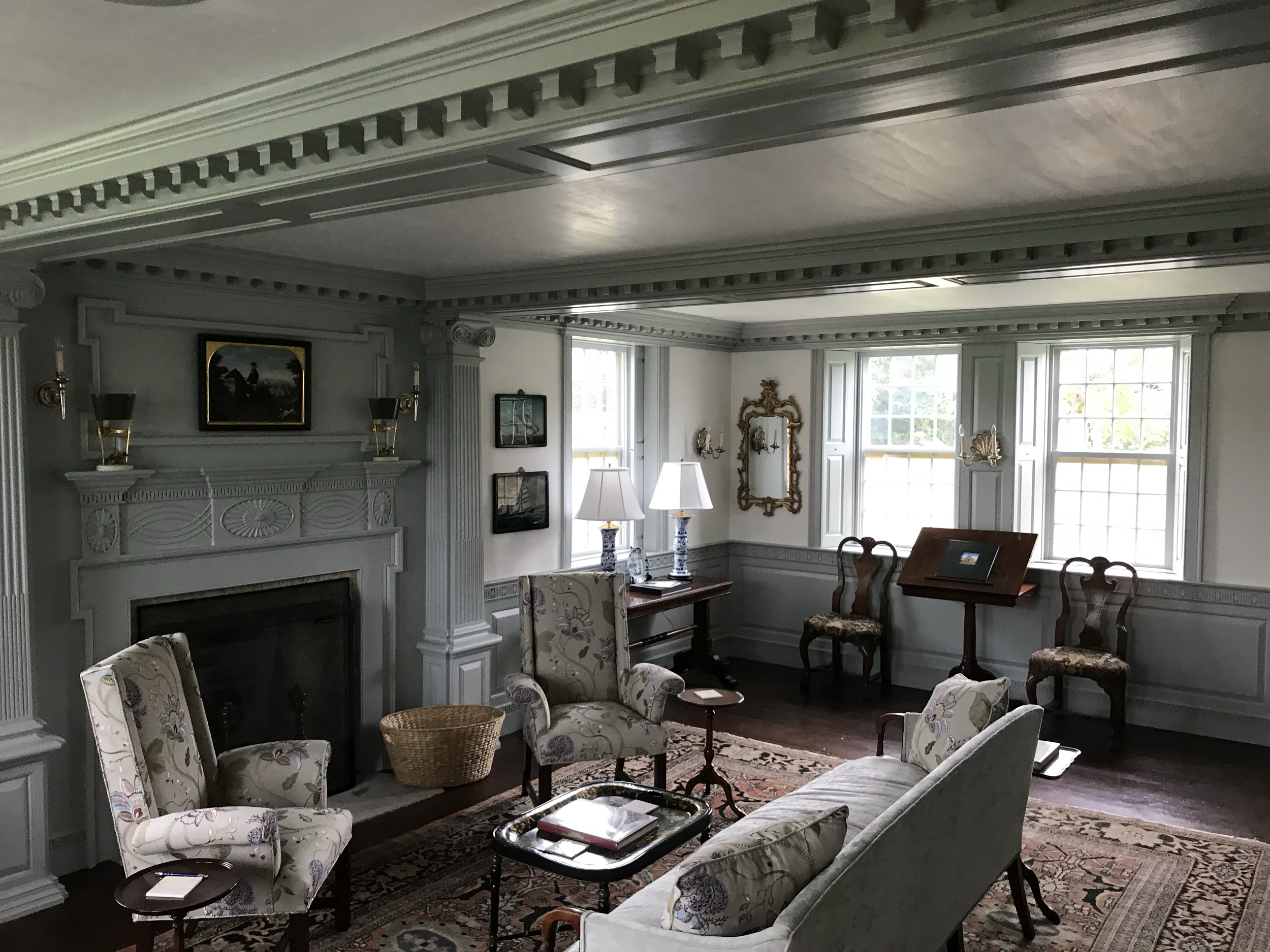A living room with gray walls and decorative crown molding on the ceiling. There are large windows with shutters, a fireplace with artwork above it, and various framed pictures on the walls. The room is furnished with patterned upholstered chairs, a 