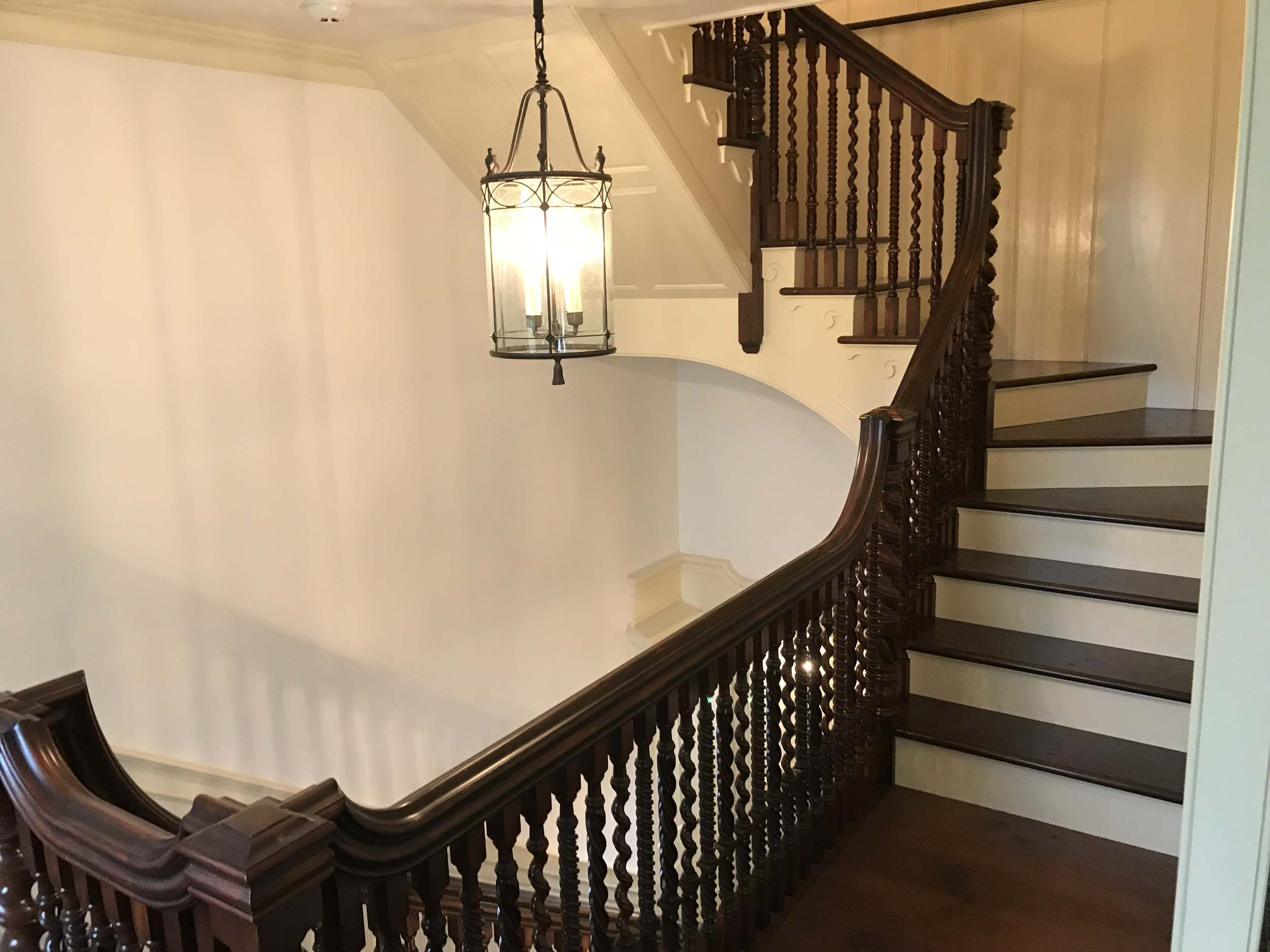 Interior view of a staircase with dark wooden handrails and spindles, a beige wall, and a chandelier hanging from the ceiling.