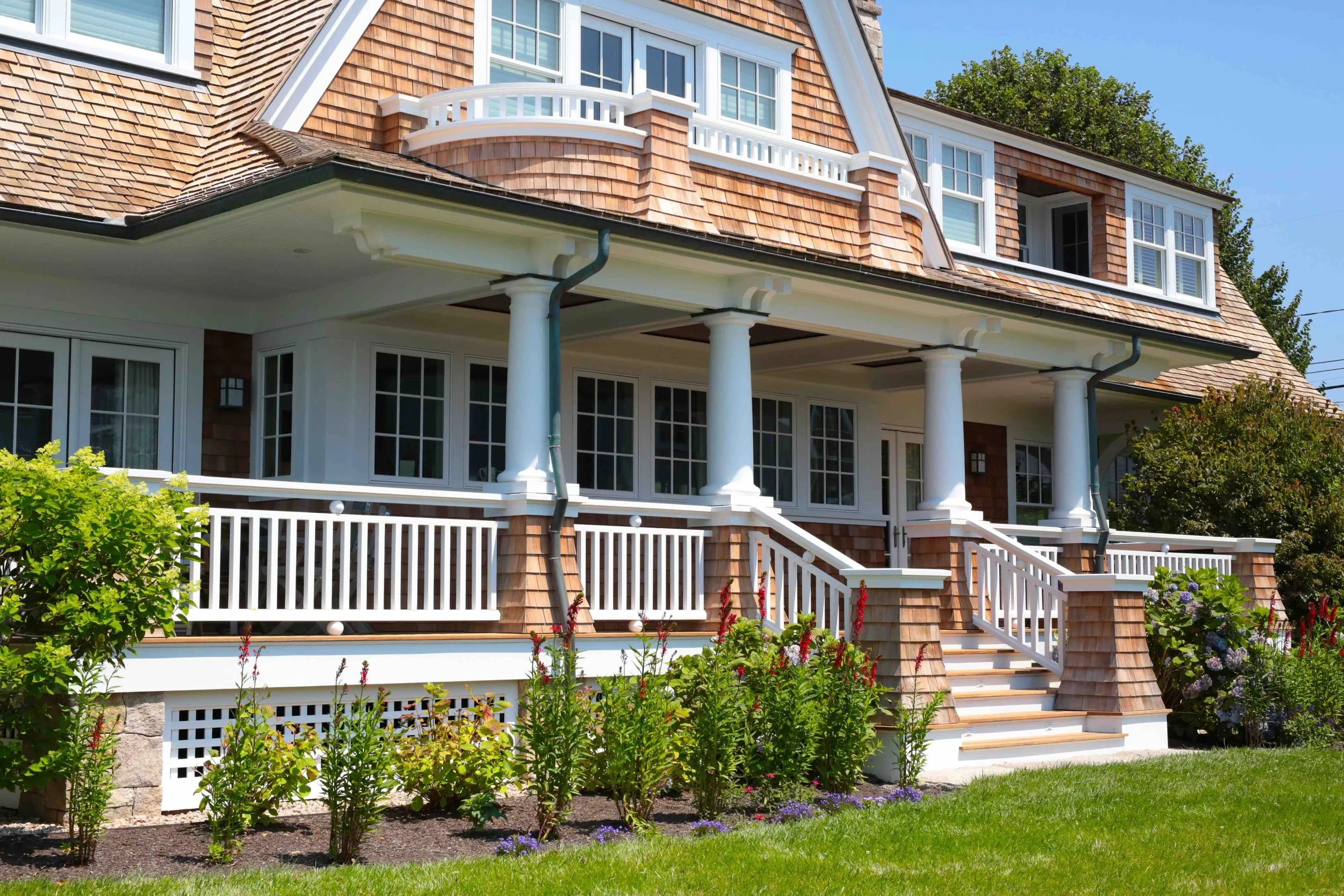 Front view of a house with wooden shingles, white columns, a balcony, and a porch, surrounded by a garden with flowers and green bushes under a clear blue sky.