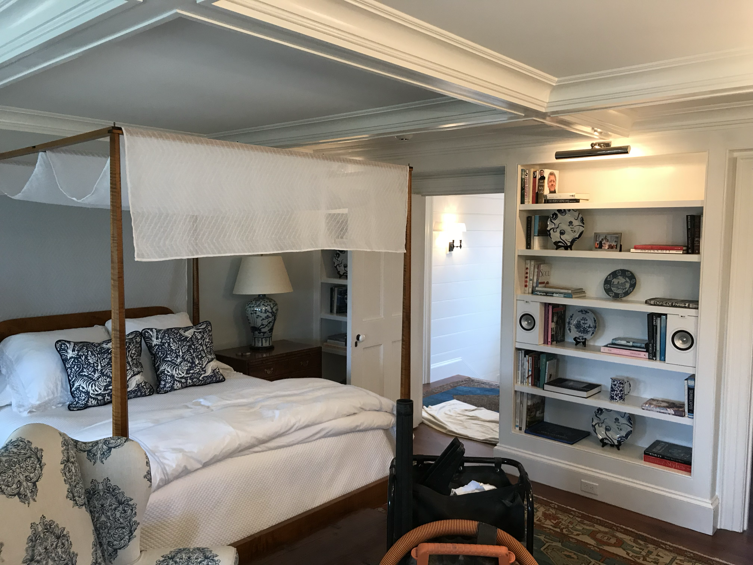 A cozy bedroom with a four-poster bed with a white mattress and dark patterned pillows. A bedside table with a ceramic lamp, a white bookshelf filled with books and decorative items, and a small rug on the wooden floor. The room has crown molding and