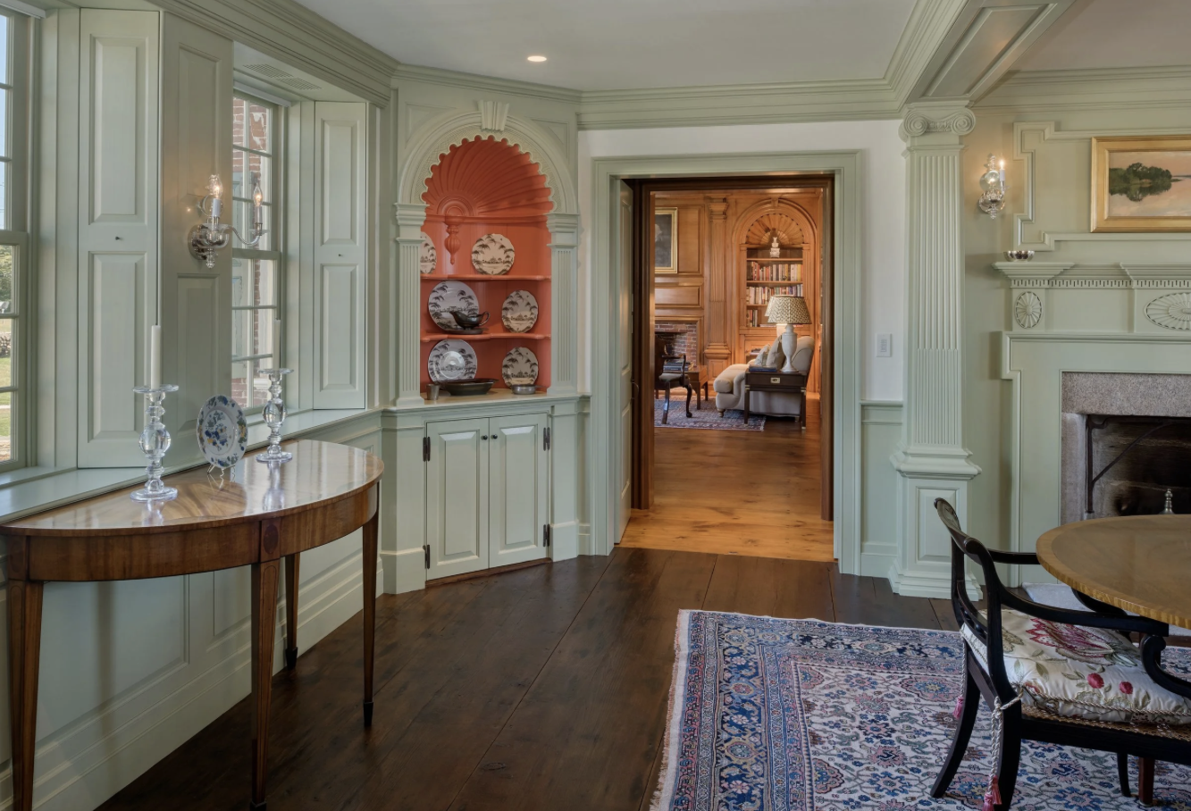 Interior view of a vintage-style living room with light green wainscoting, a built-in display cabinet with decorative plates, a dark overhanging table with candlesticks, a patterned area rug, a fireplace, and a doorway leading to another room with wo