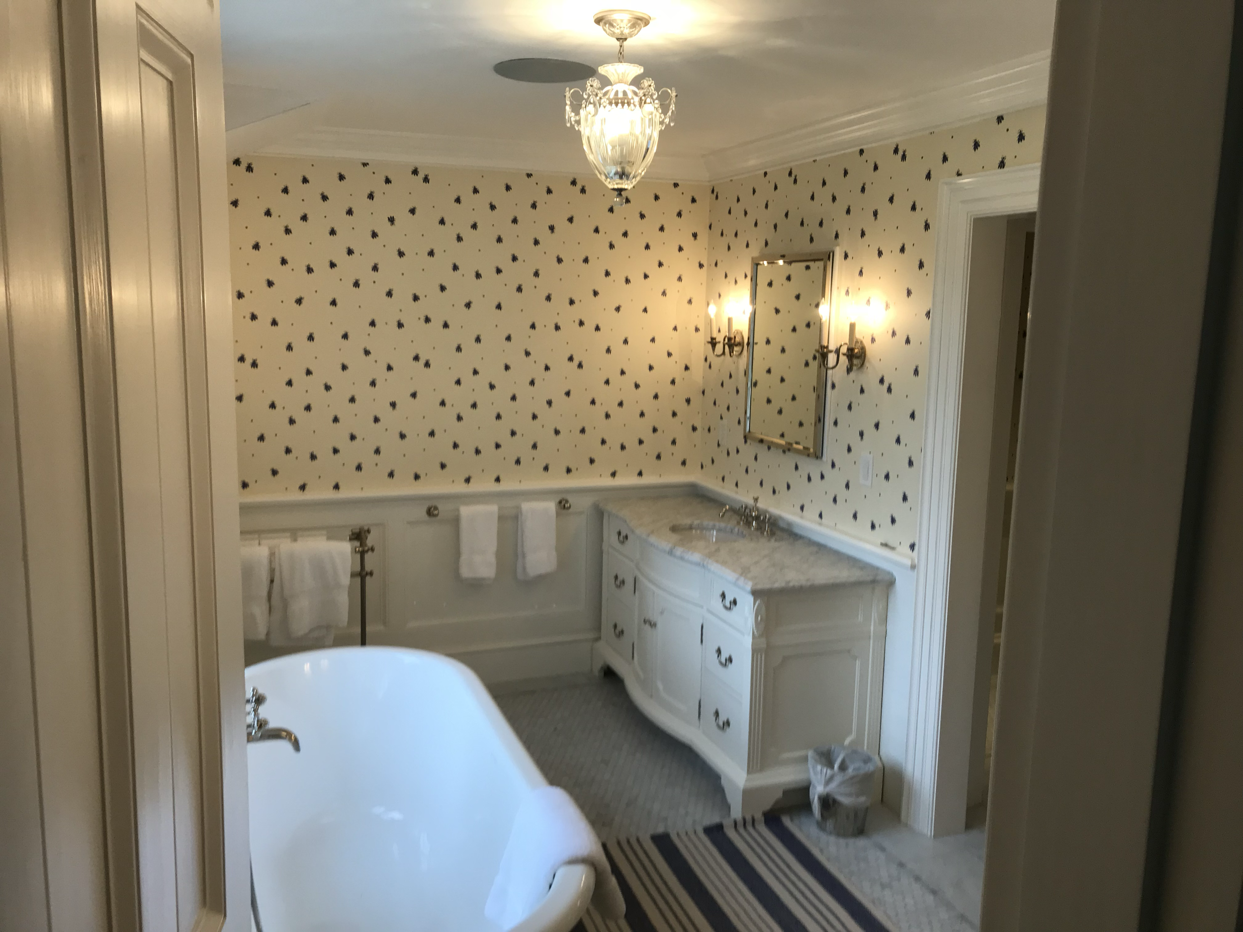 A bathroom with a clawfoot bathtub, a white vanity with multiple drawers, a large mirror, two wall-mounted sconces, a chandelier ceiling light, and wallpaper with a pattern of small black flower-like shapes on a cream background.