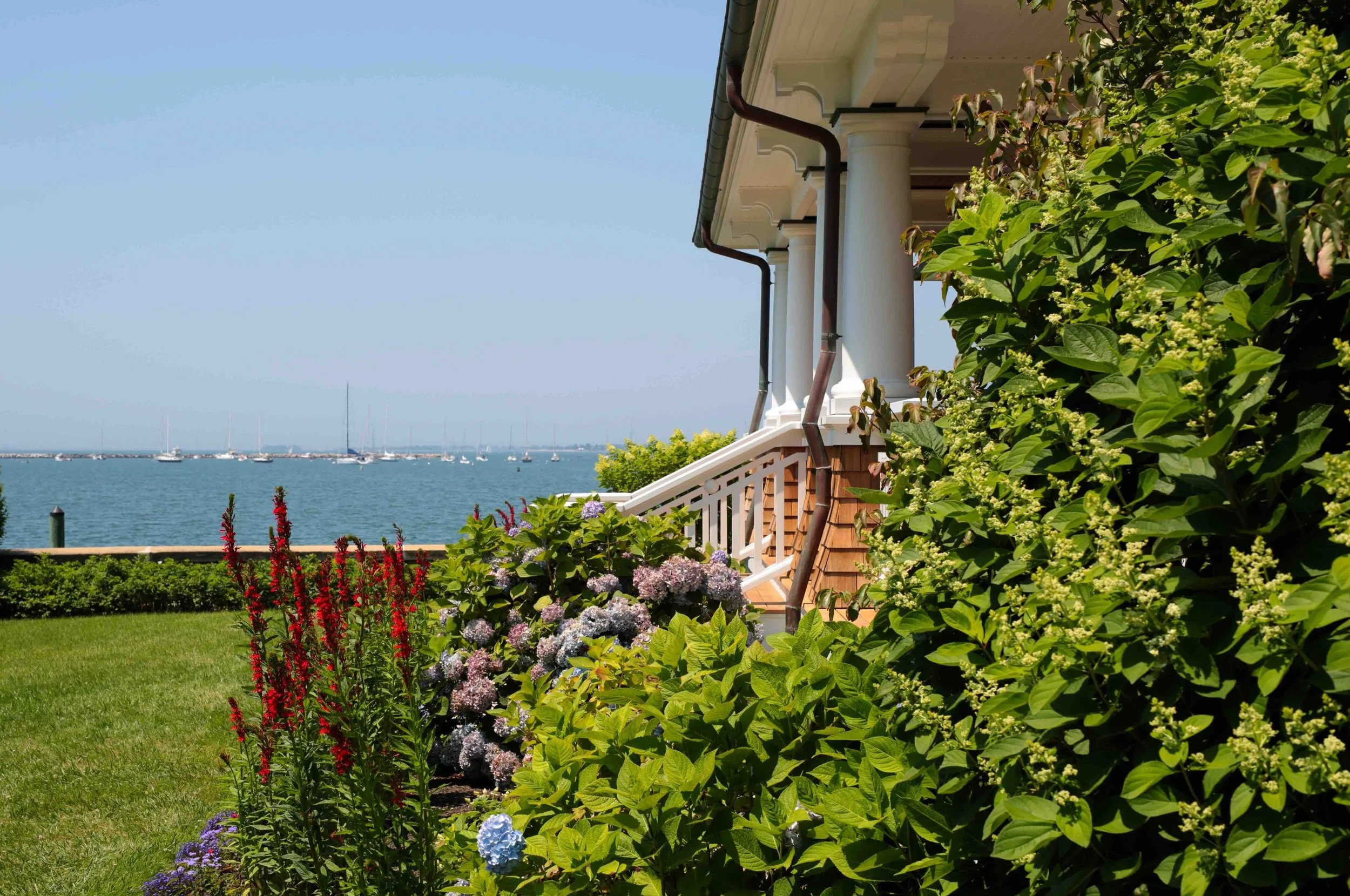 A colorful garden with blooming flowers, lush green plants, and a view of a bay with sailboats and a clear sky, next to a house with white columns and a staircase.