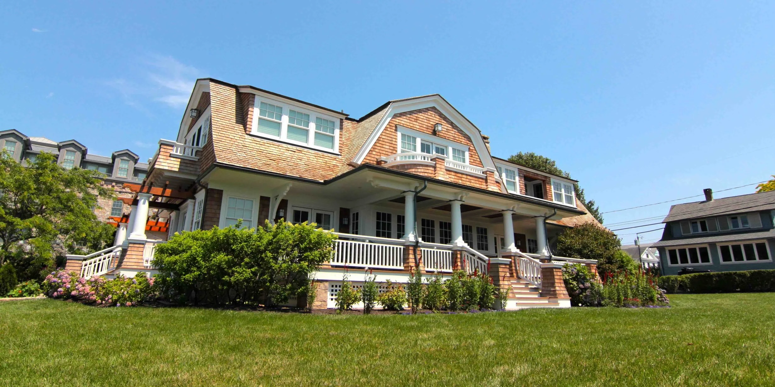 A large, multi-story house with a front porch, white railings, and a shingled roof, surrounded by a green lawn and garden under a clear blue sky.