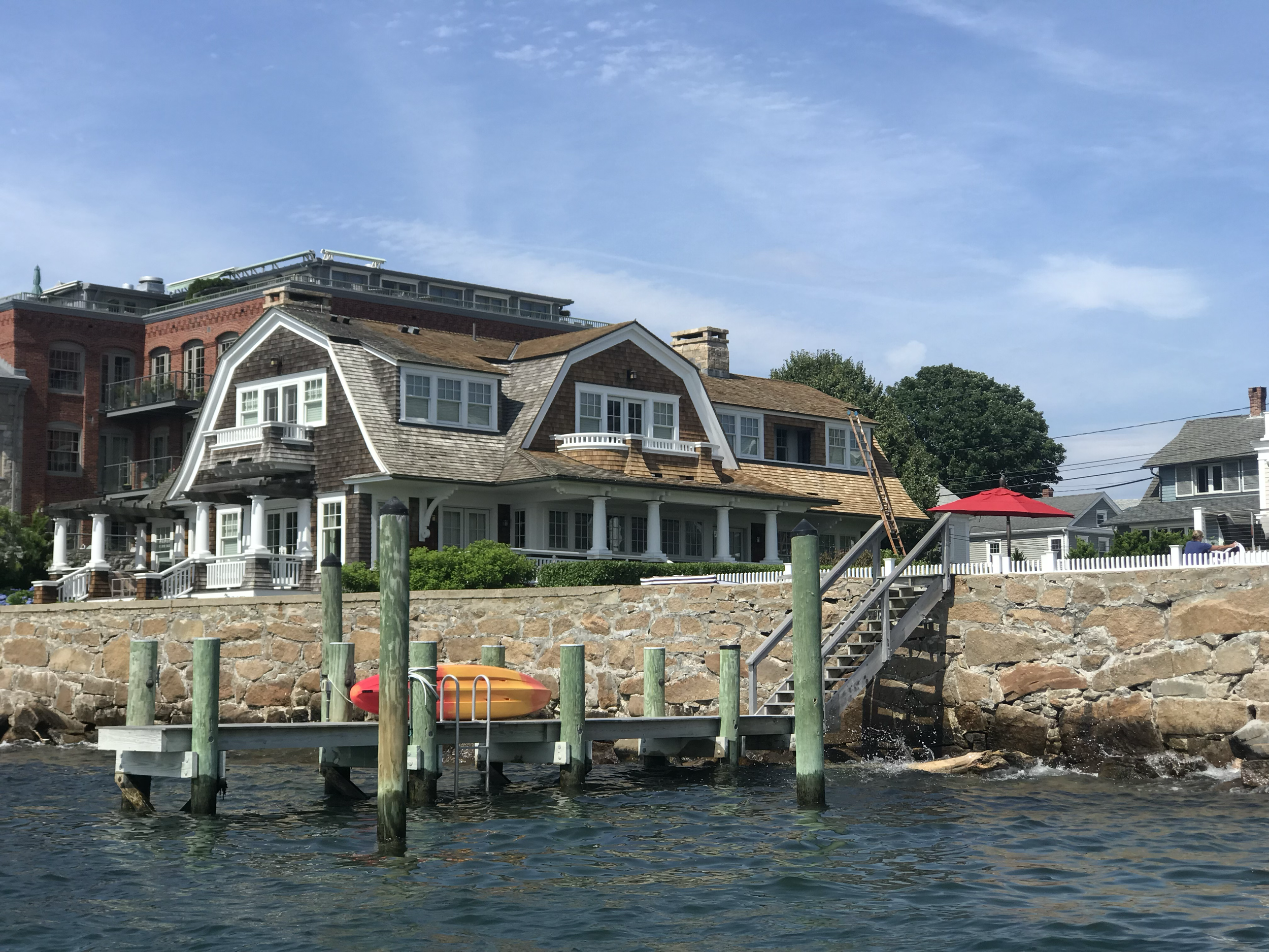 A row of residential houses by the water, with a dock and a canoe in the foreground, under a blue sky with some clouds.