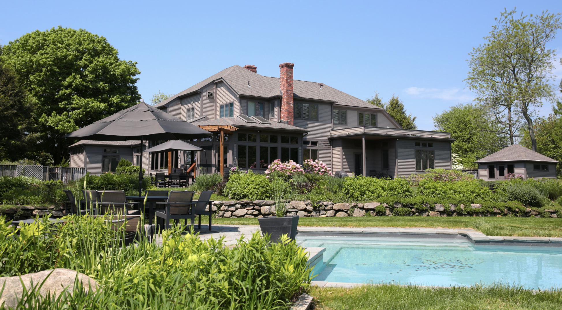 Large gray house with multiple windows, surrounded by green trees, and a landscaped backyard with patio furniture, umbrellas, and a swimming pool.