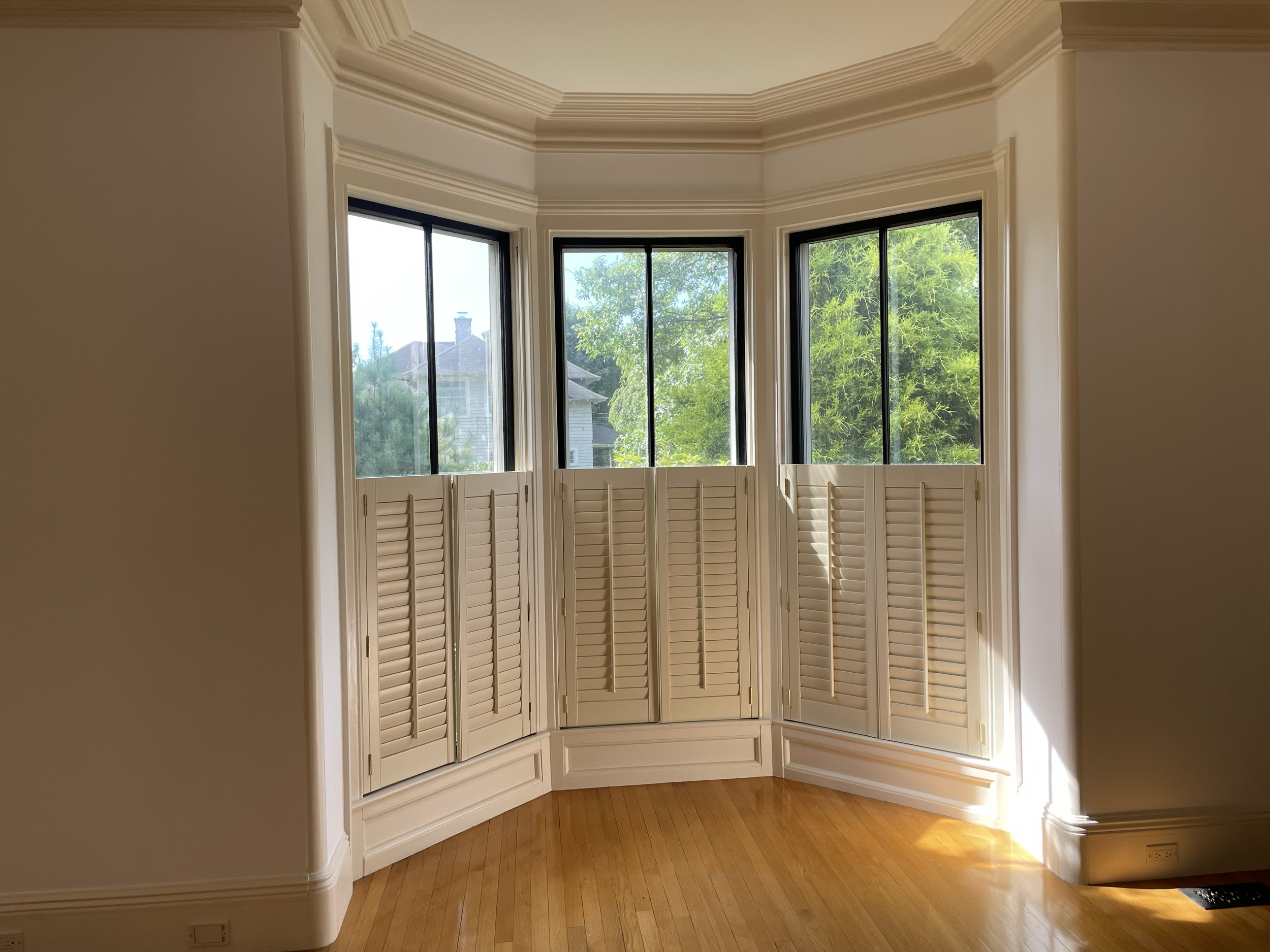 A room with a bay window with white plantation shutters, beige walls, crown molding, hardwood floors, and a view of trees and neighboring houses outside.