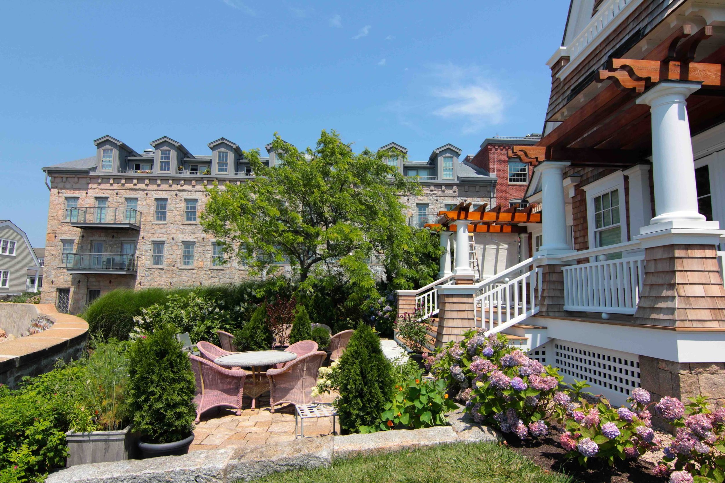 View of a residential backyard with a stone and wood house, outdoor seating area with pink chairs, blooming hydrangeas, and trees under a clear blue sky.