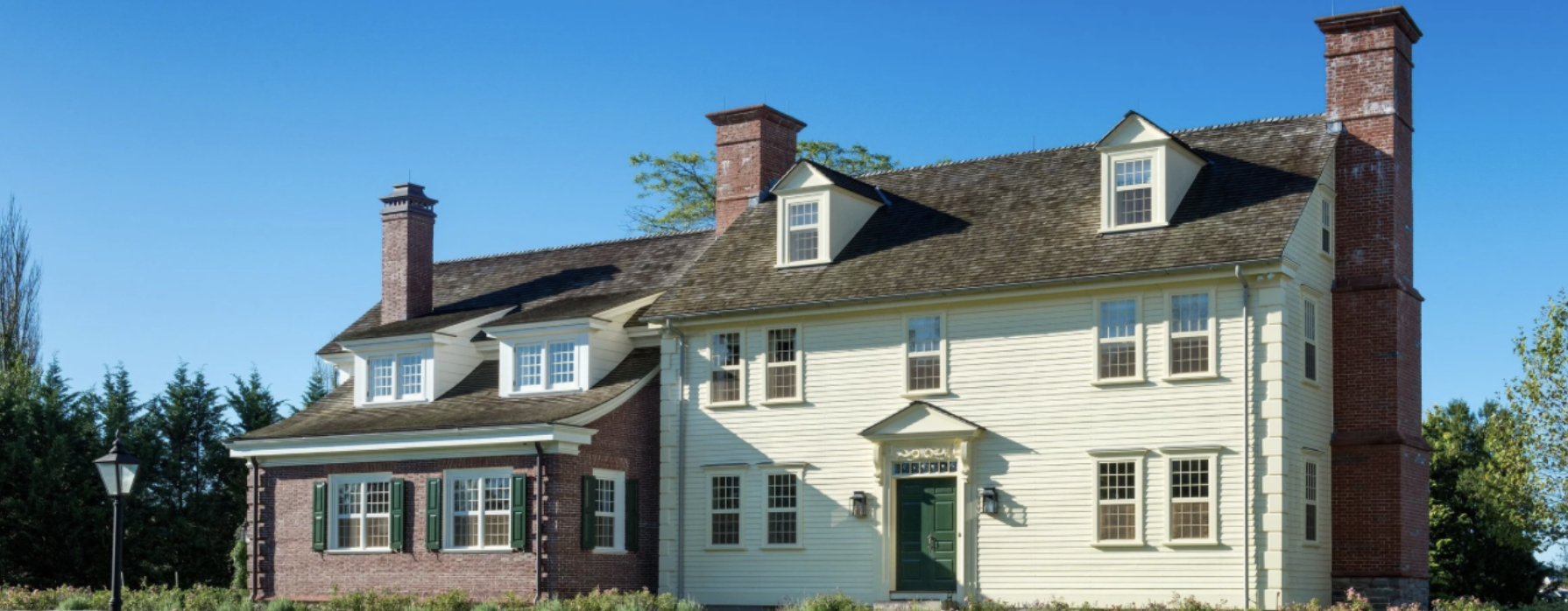 A large historic house with a mix of brick and white siding, multiple dormer windows, green shutters, and brick chimneys on a bright sunny day.