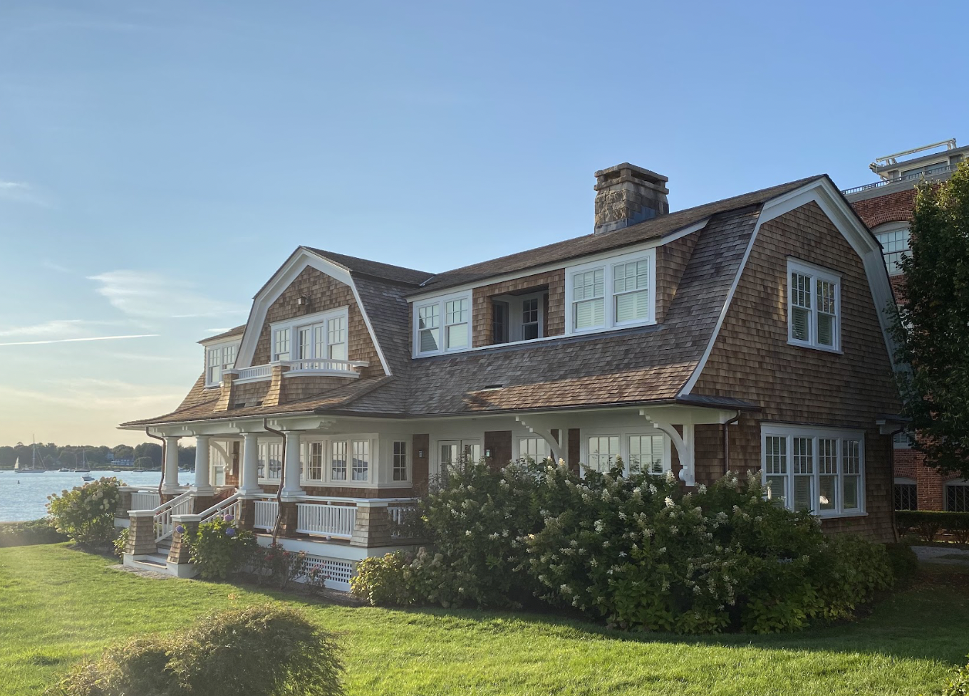 A large, two-story house with wooden shingles, white window frames, and a front porch overlooking a body of water, with lush green lawns and shrubbery.