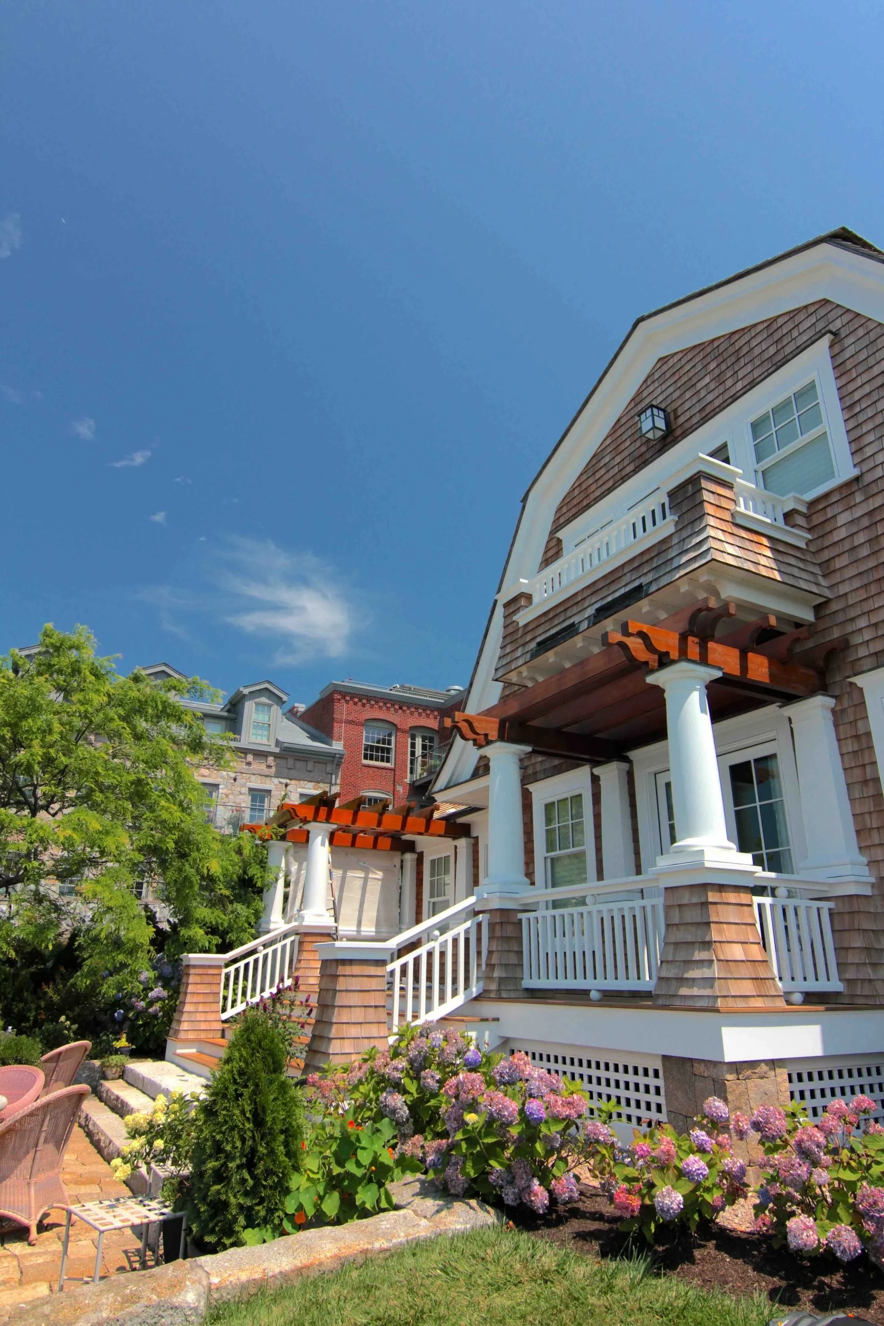 The image shows the upper part of a house with brown shingle siding, white trim, and columns supporting a small balcony. The house is set against a clear blue sky, with some trees and gardens visible at the bottom, including purple hydrangea flowers 
