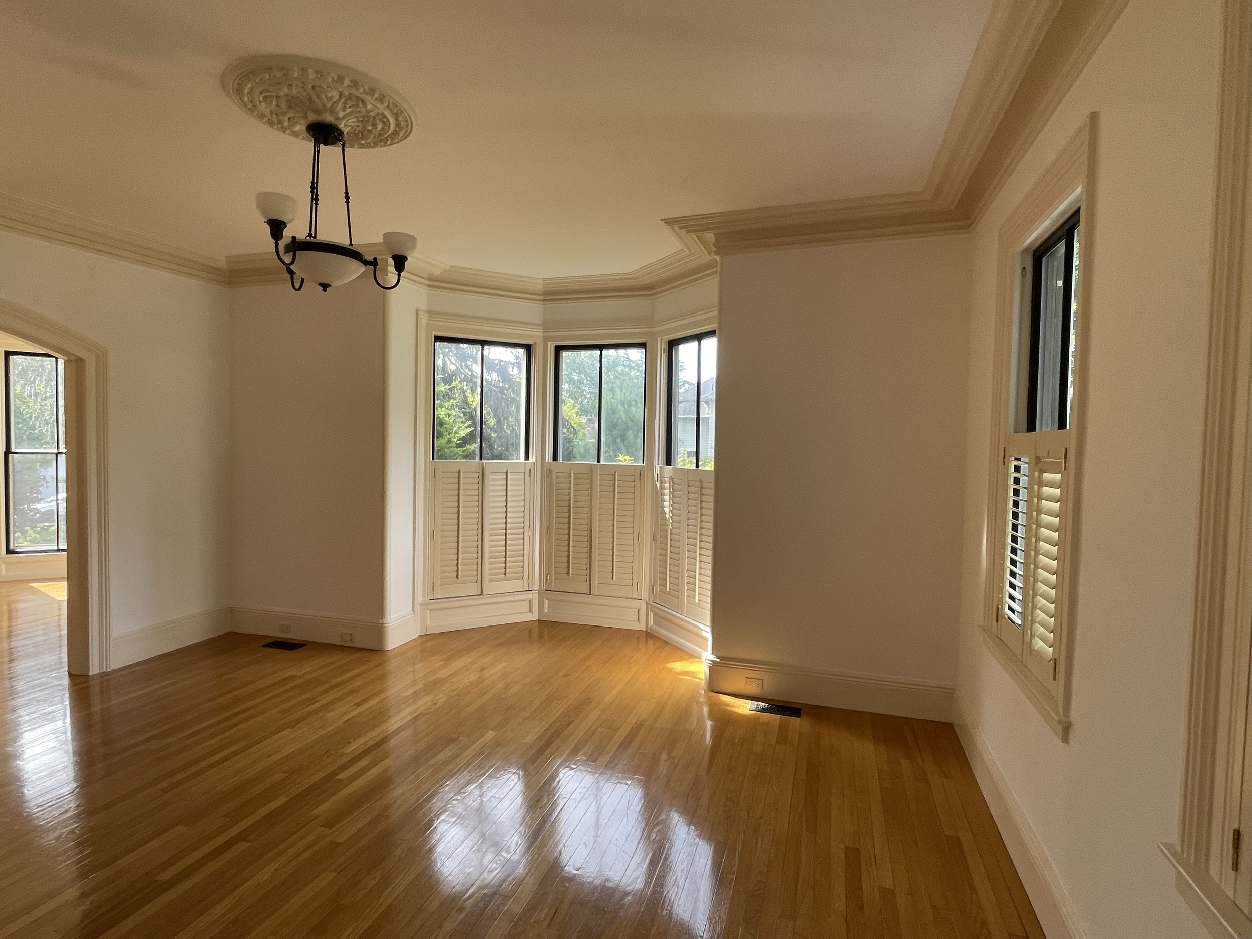 An empty living room with hardwood floors, large windows with shutters, a ceiling chandelier, and decorative crown molding.