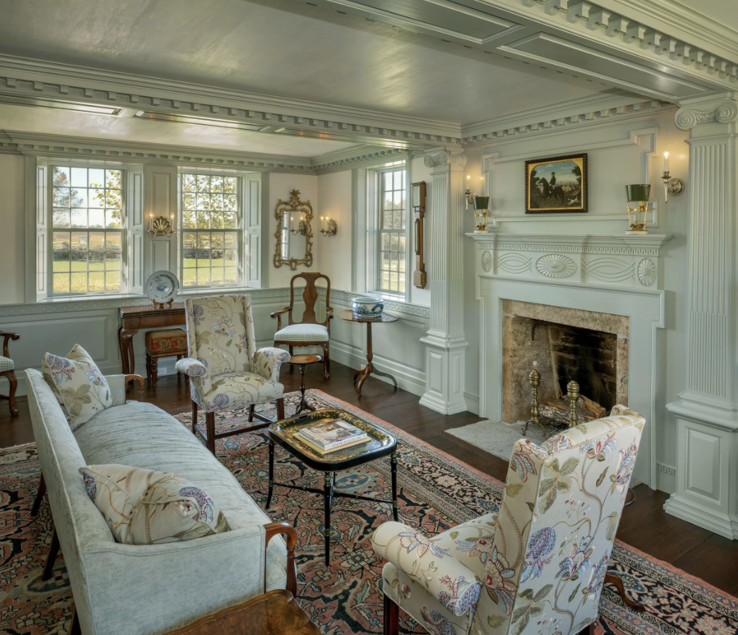 A cozy living room with vintage furniture, a fireplace, and large windows showing a sunny outdoor view.