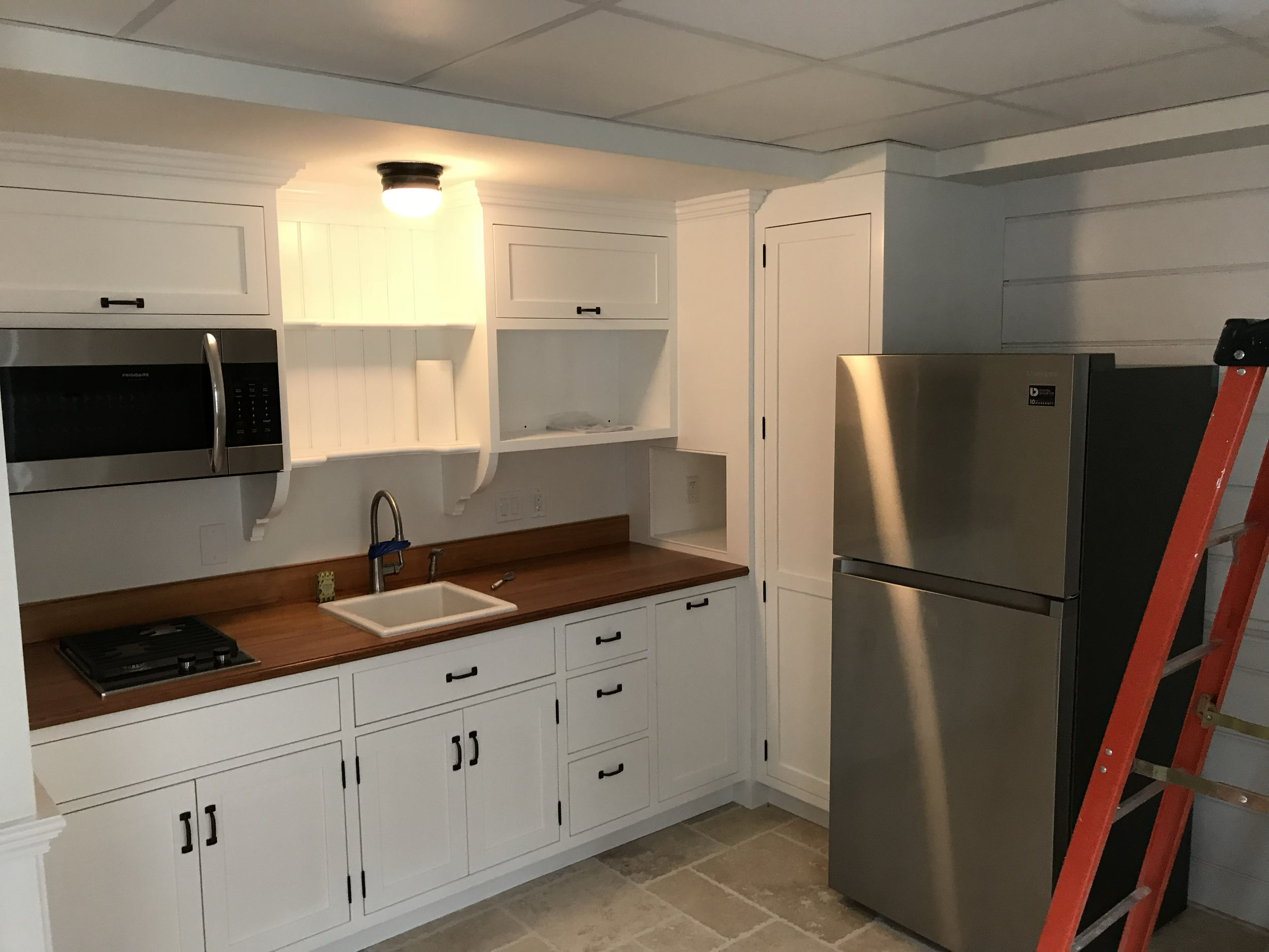 Kitchen with white cabinets, wooden countertop, stainless steel microwave, stainless steel refrigerator, and orange ladder.