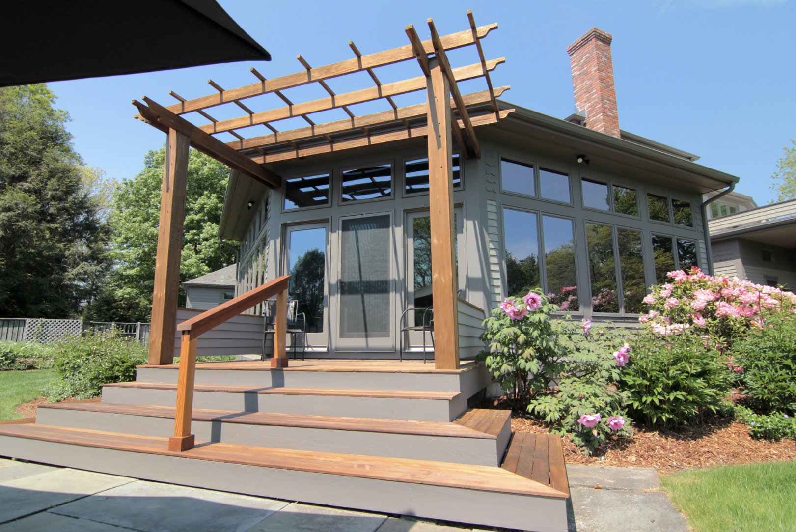 Backyard with a house featuring large windows, a sliding glass door, and a partially constructed wooden pergola over a deck, surrounded by flowering shrubs and trees.