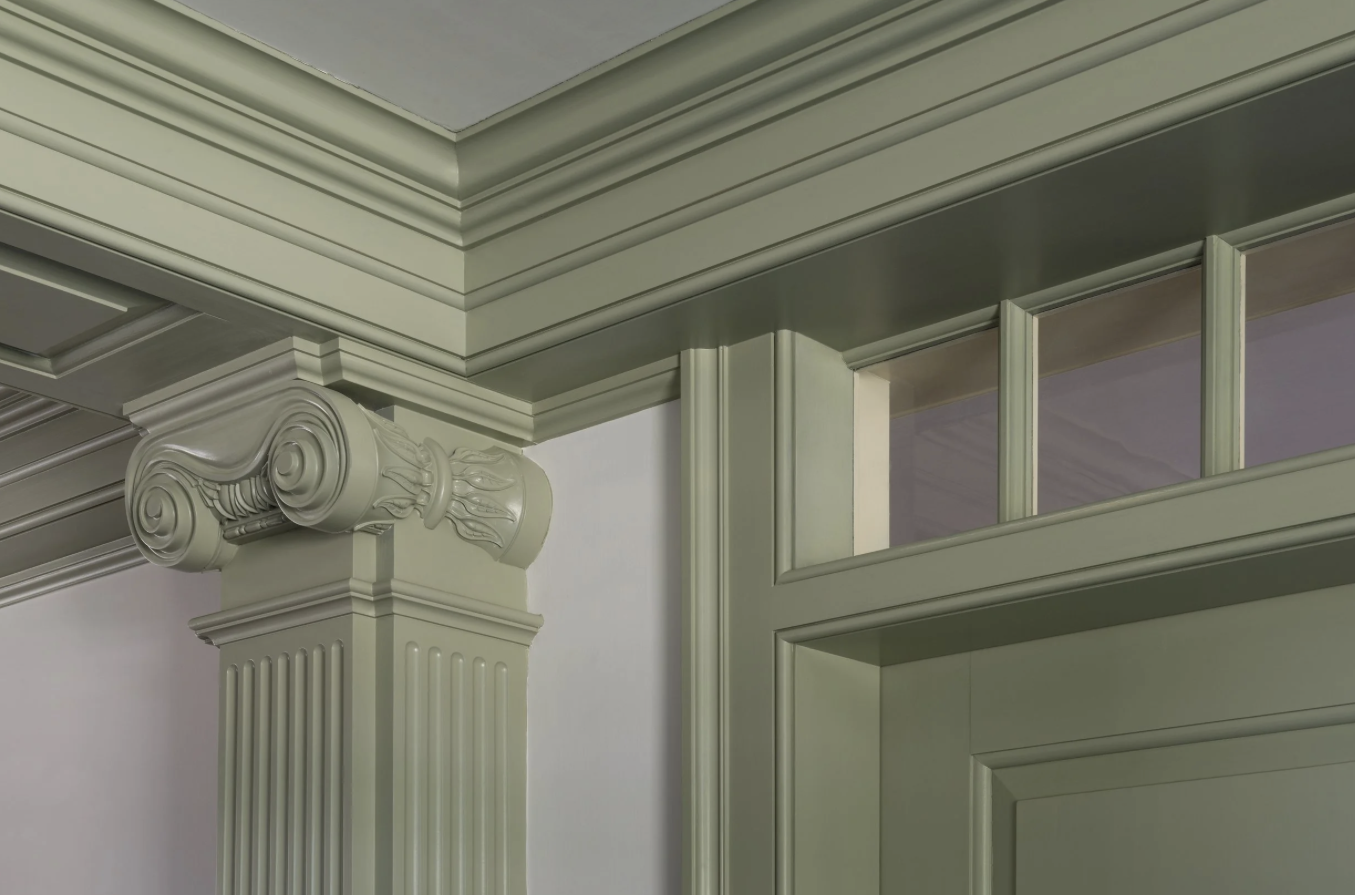 Close-up of an ornate, classical-style column with a scroll-shaped capital, painted in a light, muted green color, adjacent to a wall with detailed molding and paneling.