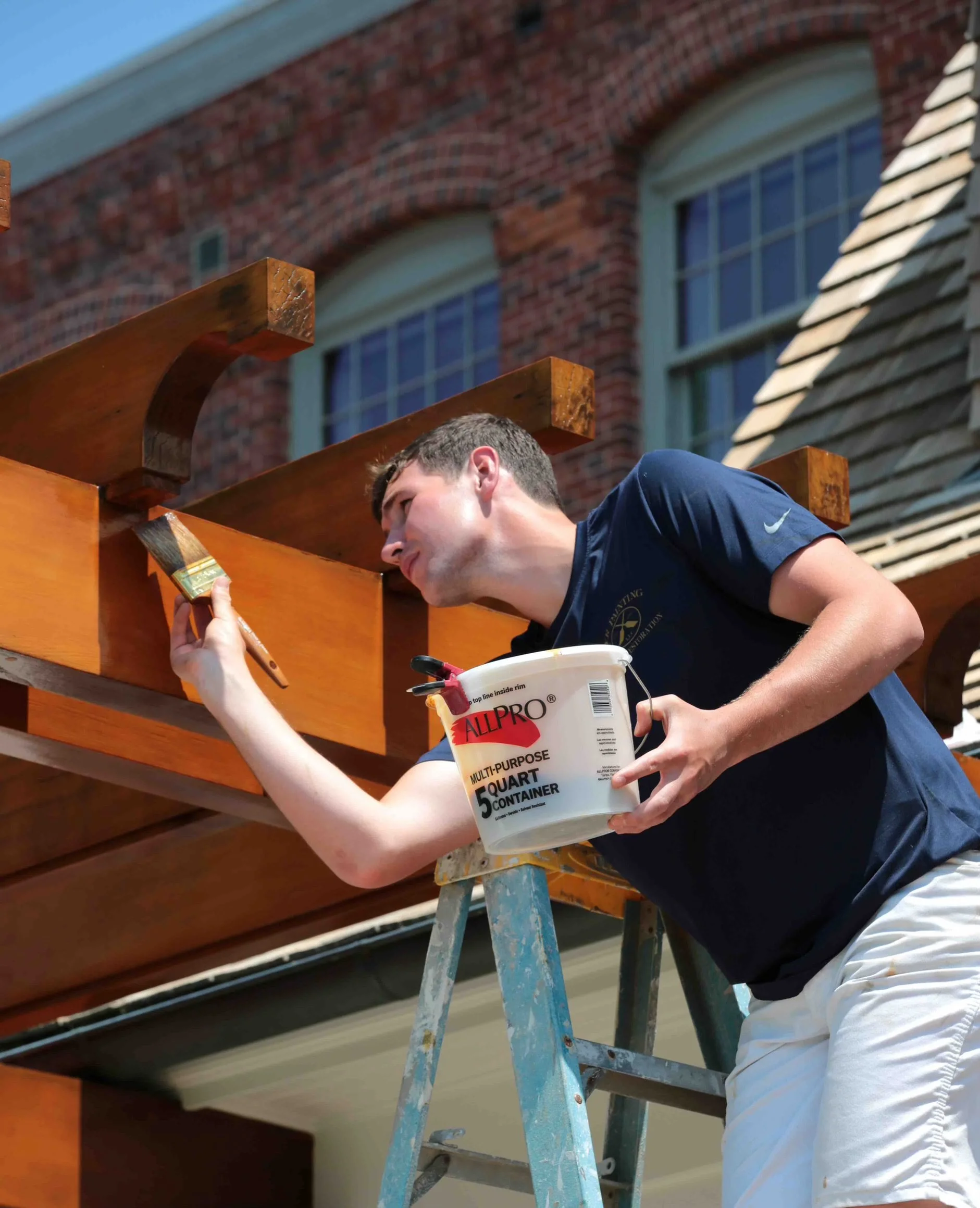 A man painting a wooden structure outdoors, standing on a ladder, with a brick building and a window in the background. He is holding a paintbrush and a bucket of paint.