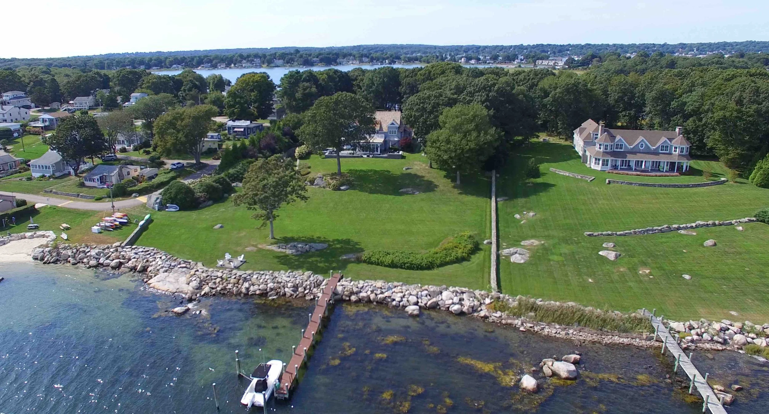 Aerial view of a lakeside neighborhood with large houses, green lawns, trees, a pier extending into the water, and a shoreline with rocks.
