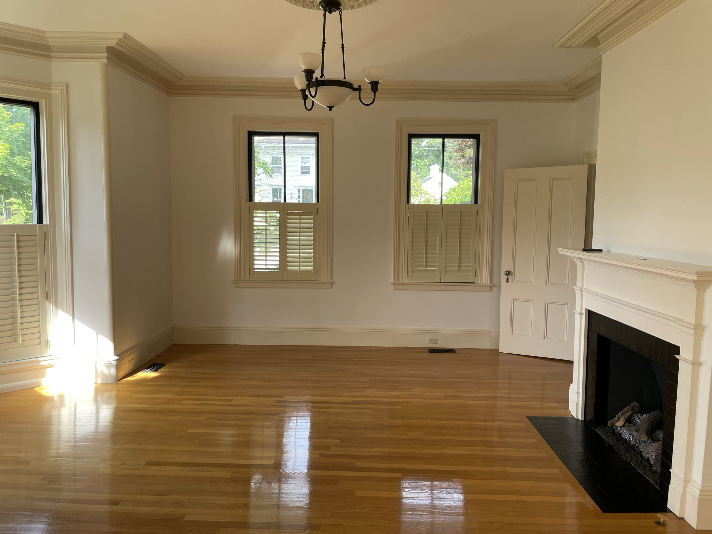 Empty living room with large windows, hardwood floors, white walls, and a white fireplace with a black interior.