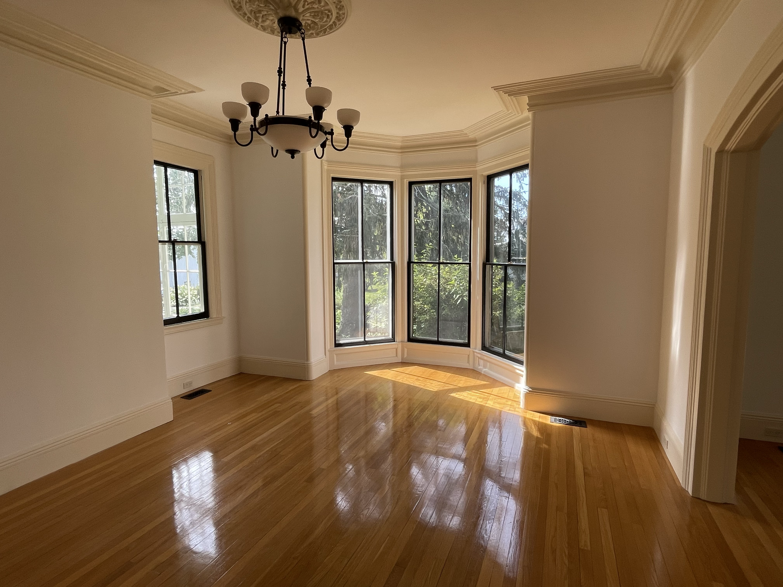 An empty room with hardwood floors, beige walls, crown molding, and large windows overlooking greenery. A black chandelier hangs from the ceiling.