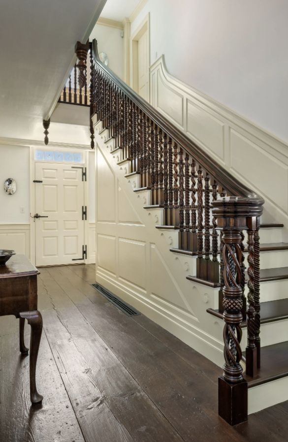Interior view of a vintage staircase with carved dark wood railings and spindles, white paneled walls, and a wooden floor. A closed door with a frosted glass window is at the top of the stairs.