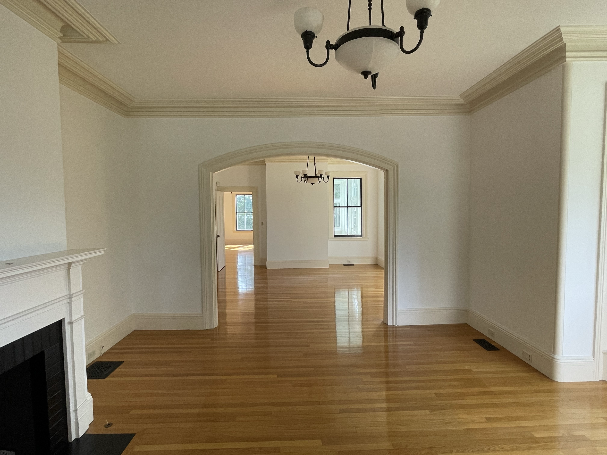 Interior view of a house showing multiple rooms with hardwood floors, white walls, crown molding, and black chandeliers. There are windows allowing natural light into the rooms.
