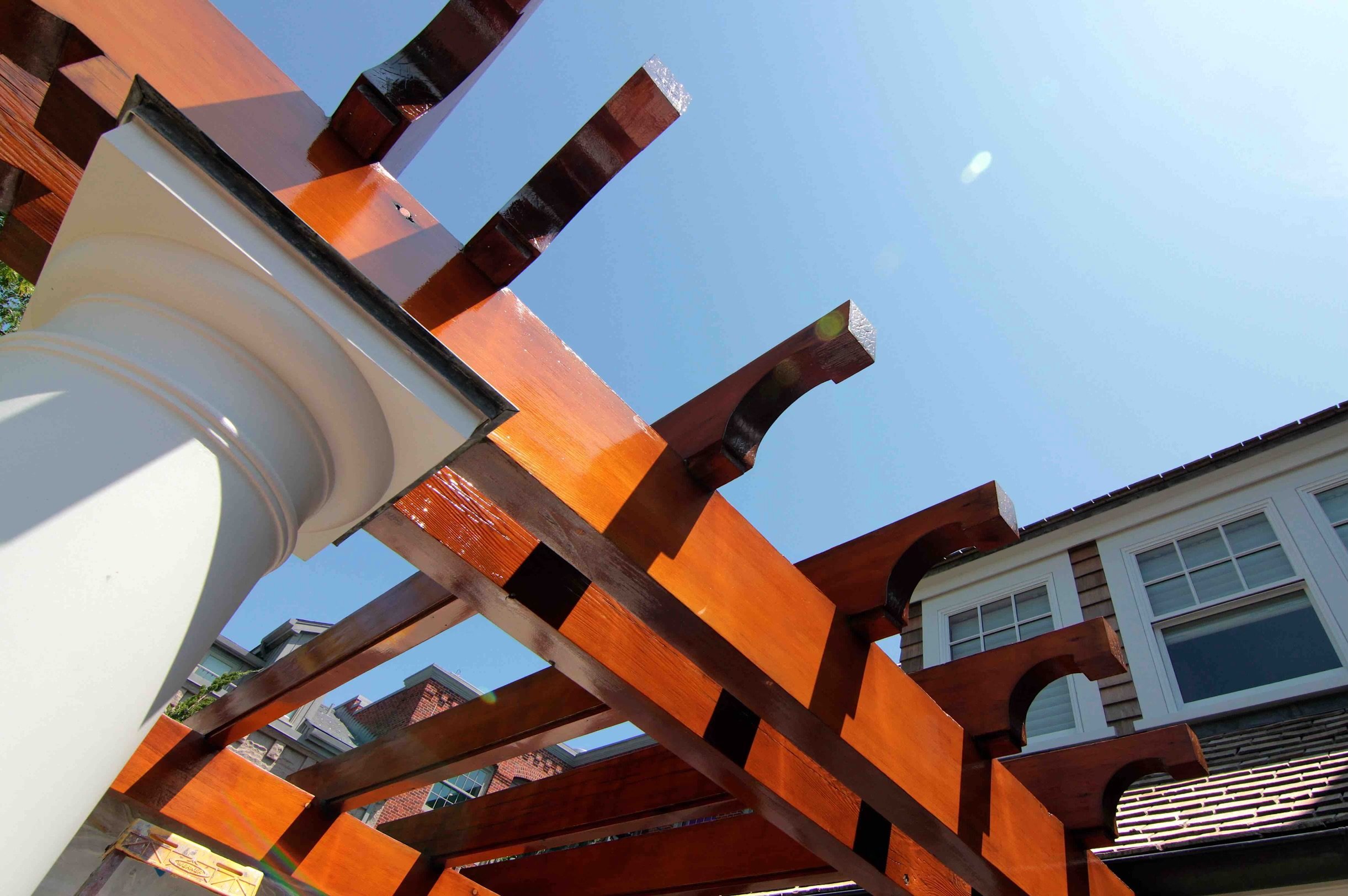 Close-up of a wooden pergola with a white gutter, under a bright blue sky with sunlight.