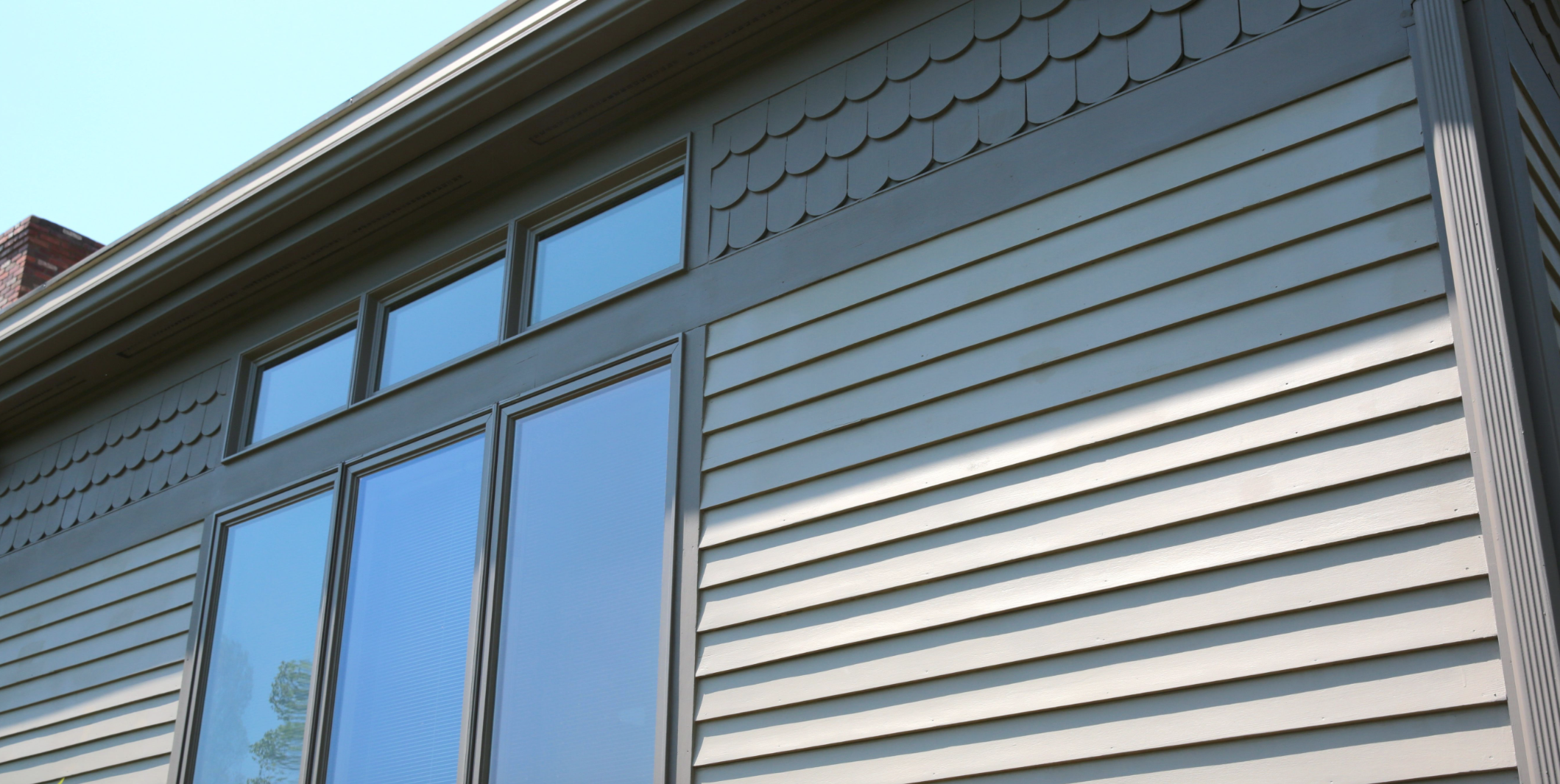 Close-up of a house's exterior showing three large windows with blue glass, horizontal cream-colored vinyl siding, scalloped shingle-style siding above the windows, and a clear sky in the background.