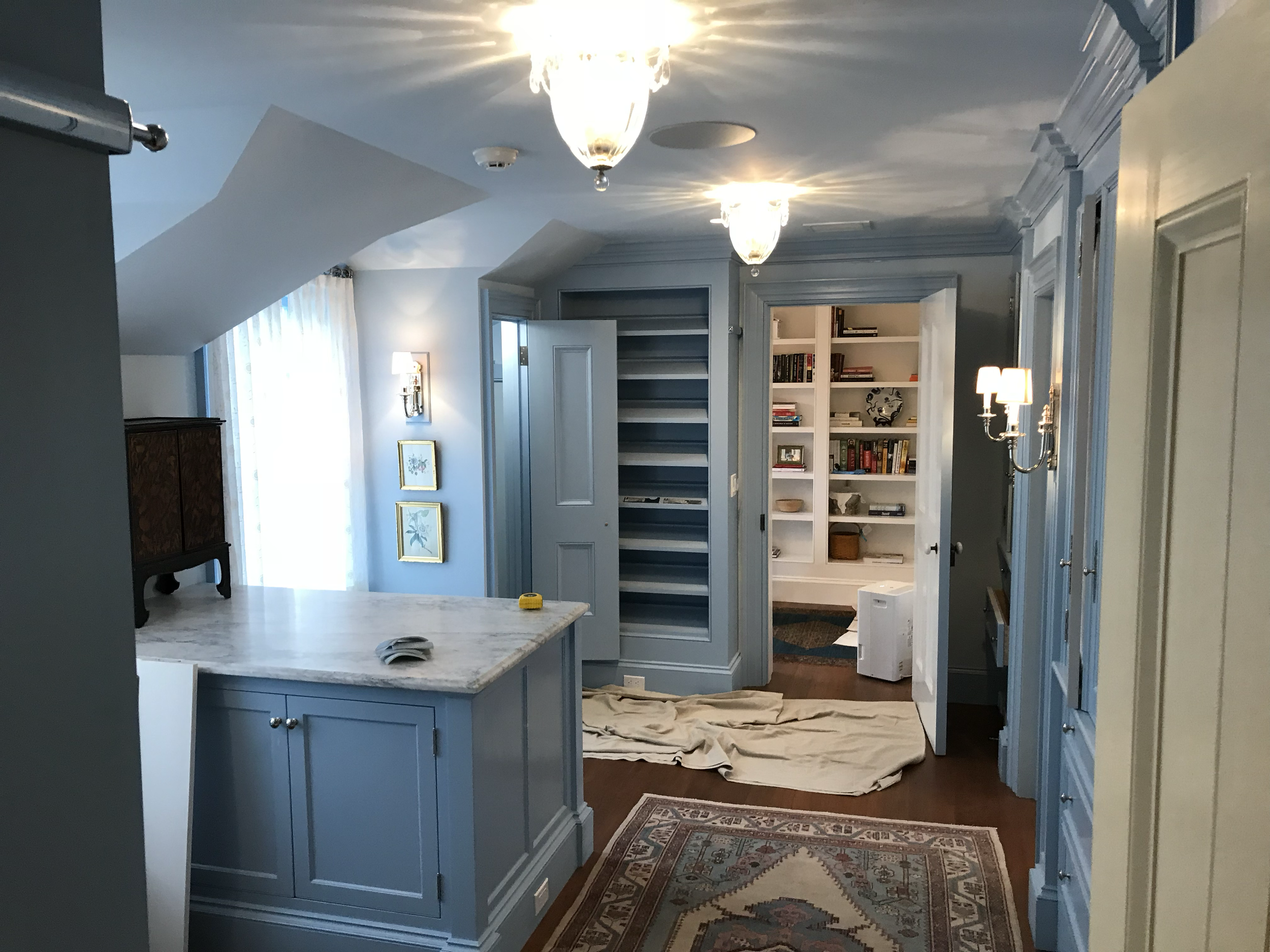 A kitchen under renovation with a drop cloth on the floor, an open cabinet, and a marble countertop. There are two chandeliers and wall sconces lighting the room, with shelves and books in the background.