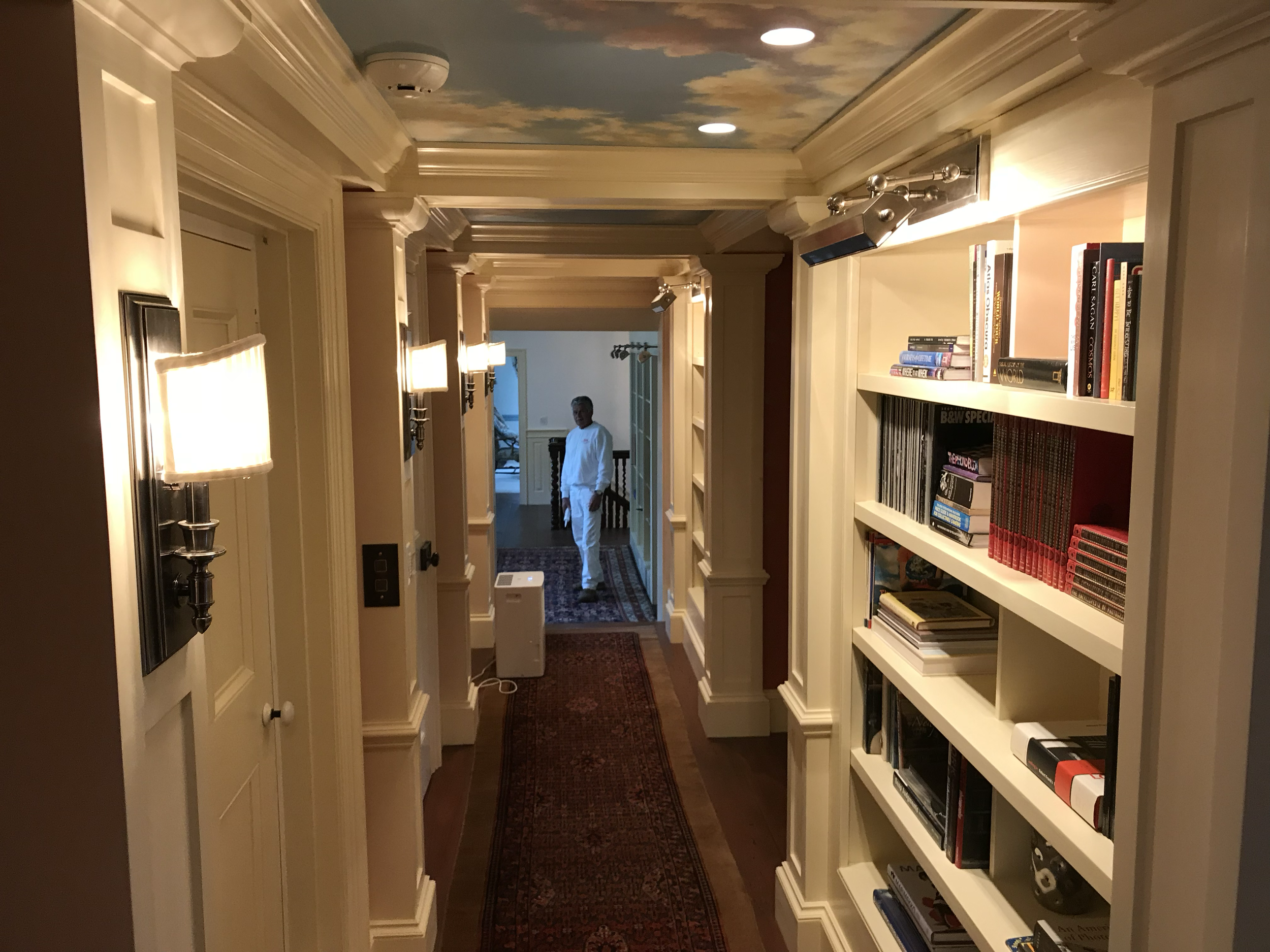 A hallway in a house with cream-colored bookshelf on the right filled with books and a red-and-black patterned rug on the wooden floor. The ceiling has a sky mural with clouds, and there are wall-mounted lights providing warm lighting. A person dress