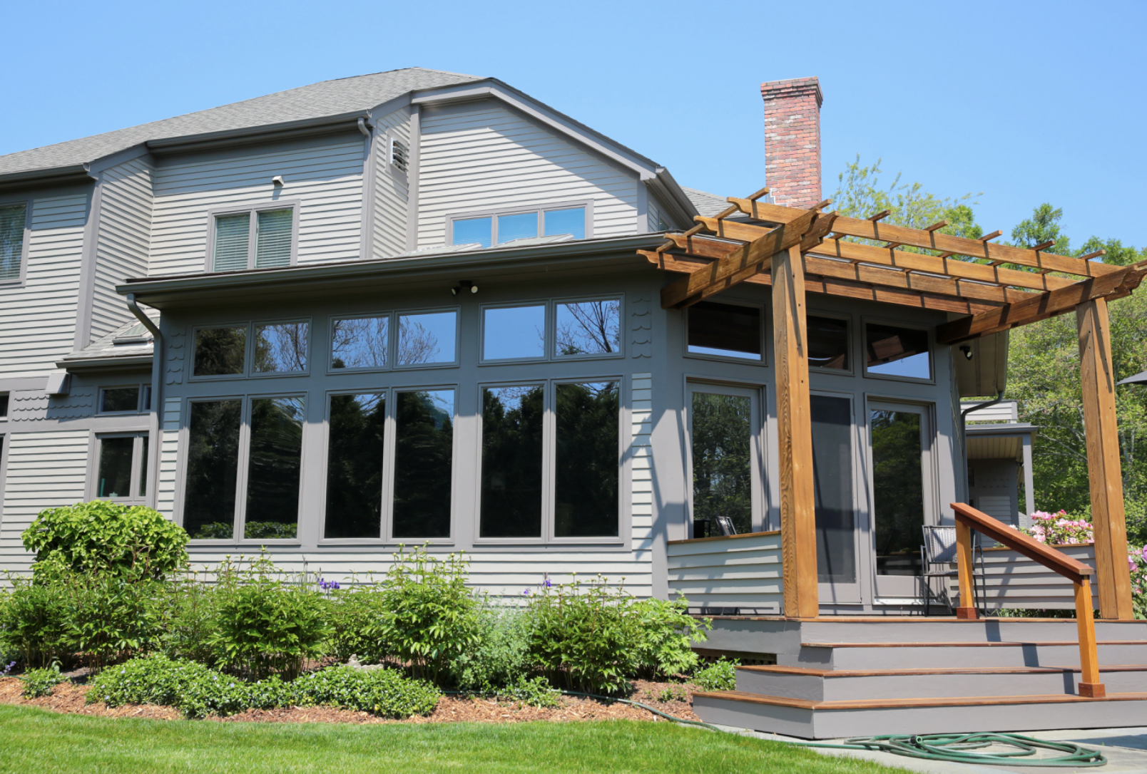 Front view of a two-story house with gray siding, large sunroom with windows, and a wooden pergola on the deck, under a clear blue sky.