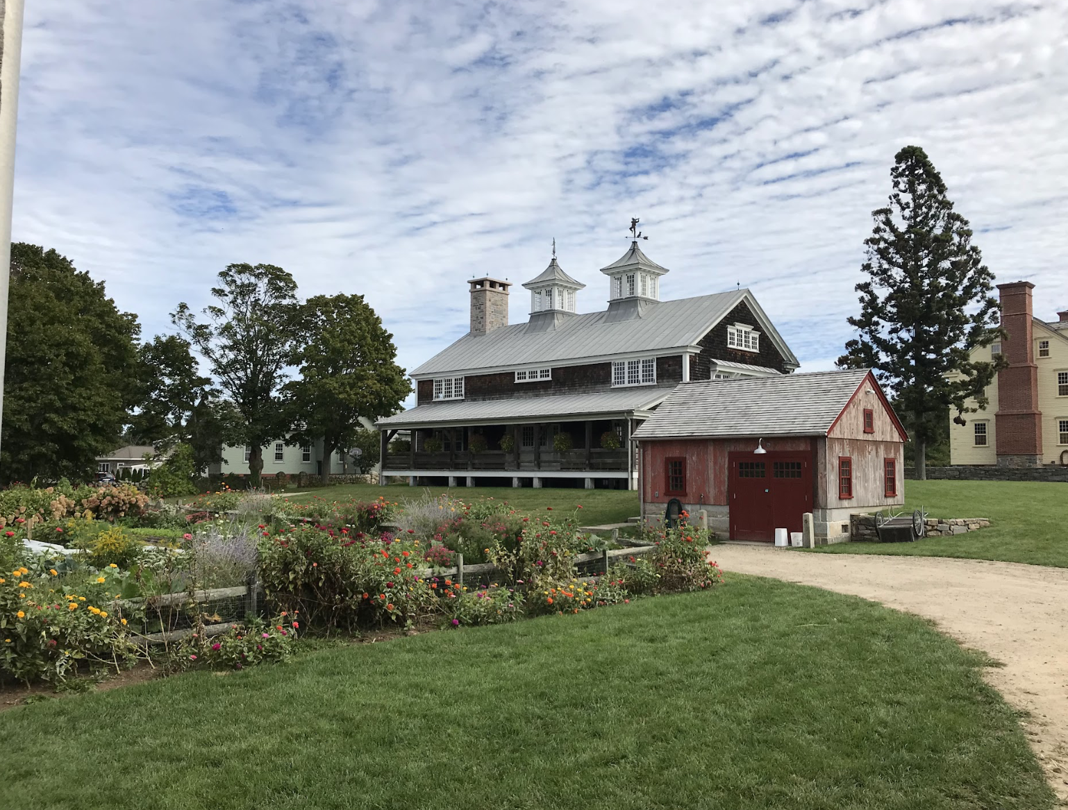 A historic barn with Patina siding and a metal roof, surrounded by colorful flowers and a well-kept lawn, with mature trees and a partly cloudy sky in the background.