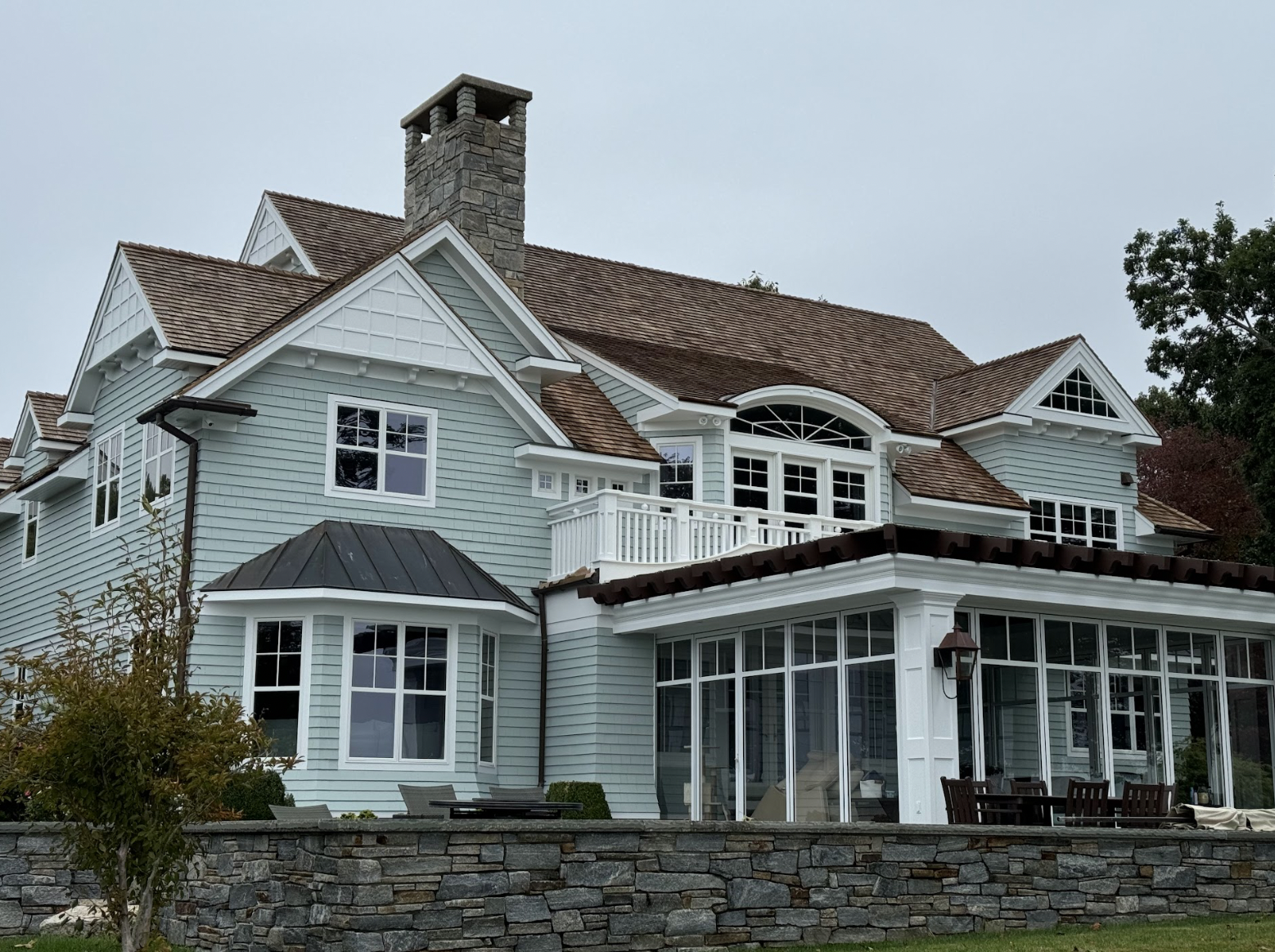 Large, light blue, multi-story house with a stone chimney, gable roofs, and various windows, including a sunroom and balcony, surrounded by trees and a stone wall.