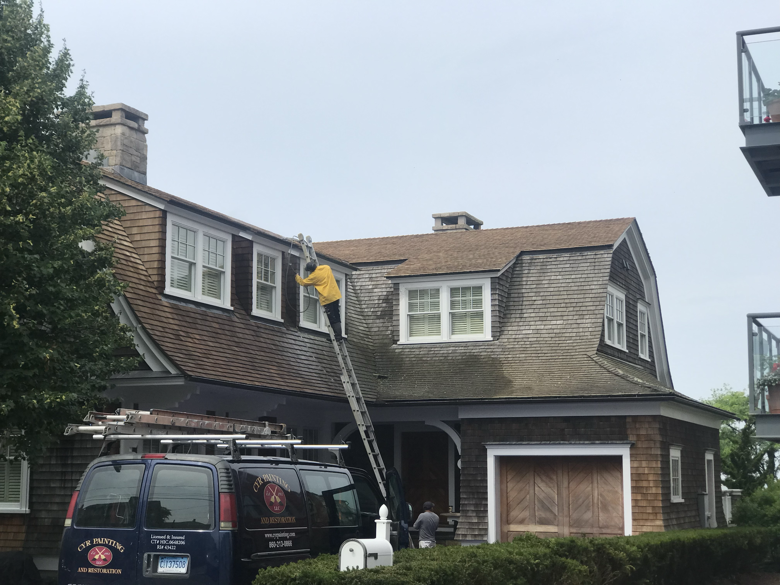 A person in a yellow jacket is standing on a ladder cleaning the roof of a house. The house has a shingled roof with multiple windows, and a tree is visible on the left. A black van with company branding is parked in front of the house. The sky is ov
