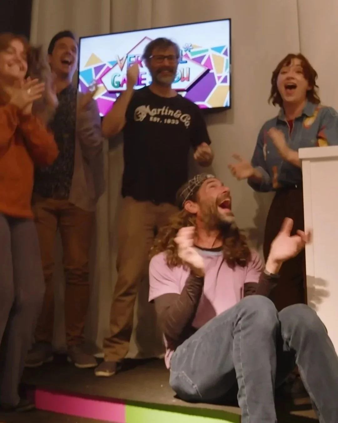 Group of six people, four women and two men, celebrating and laughing together indoors.