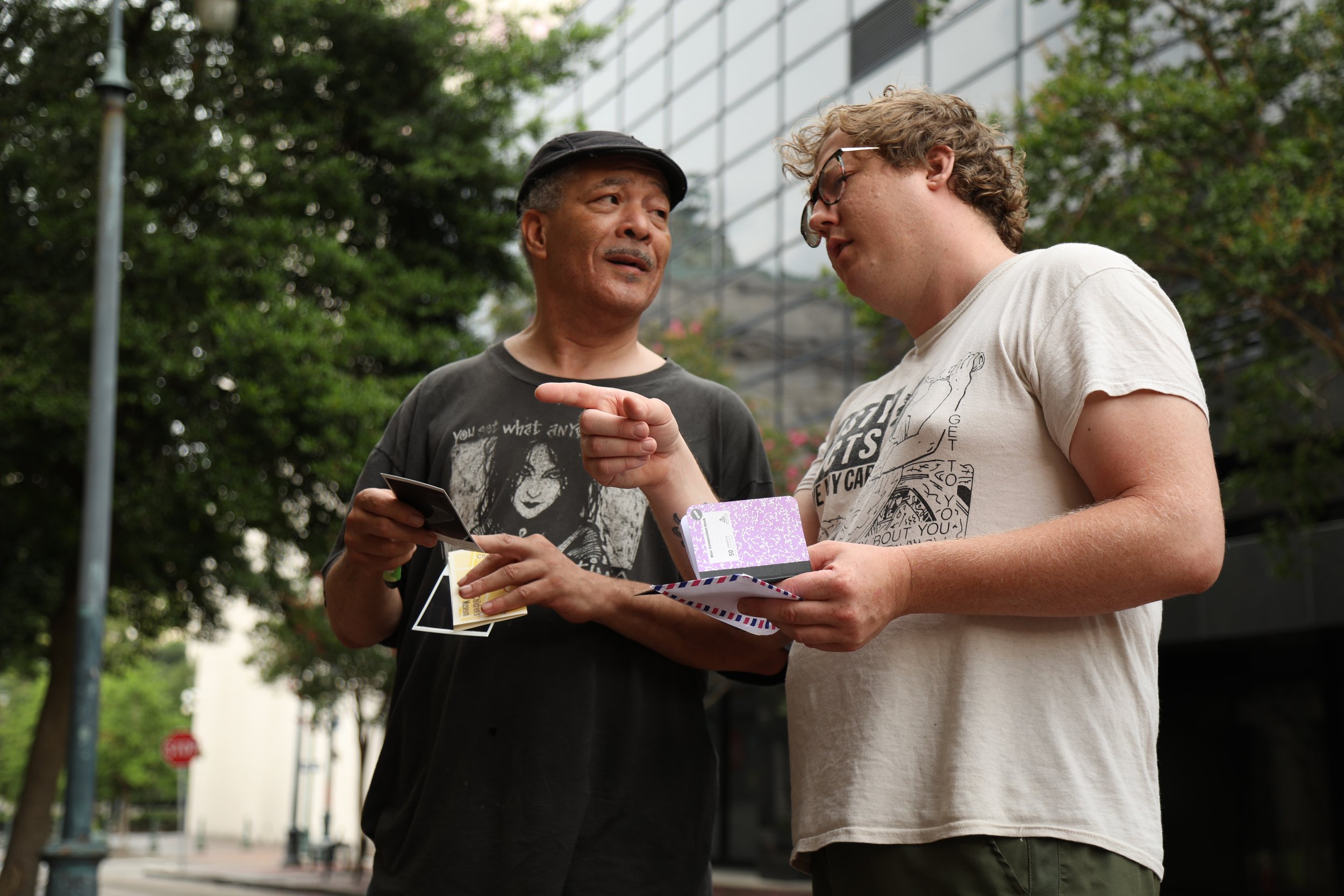 Two men are having a conversation outdoors in a park with trees and a modern building in the background. One man is gesturing with his finger while holding papers and a card. The other man is listening and holding papers and a card.