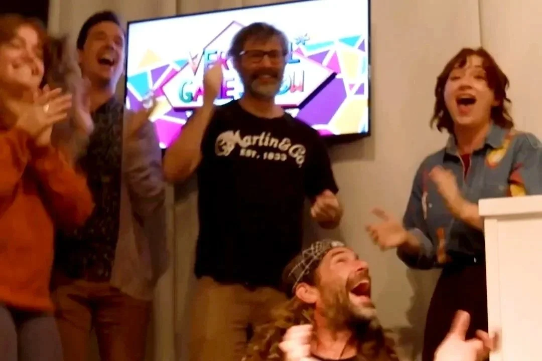 Group of five people celebrating and cheering with smiles and laughter, standing in front of a colorful TV screen.