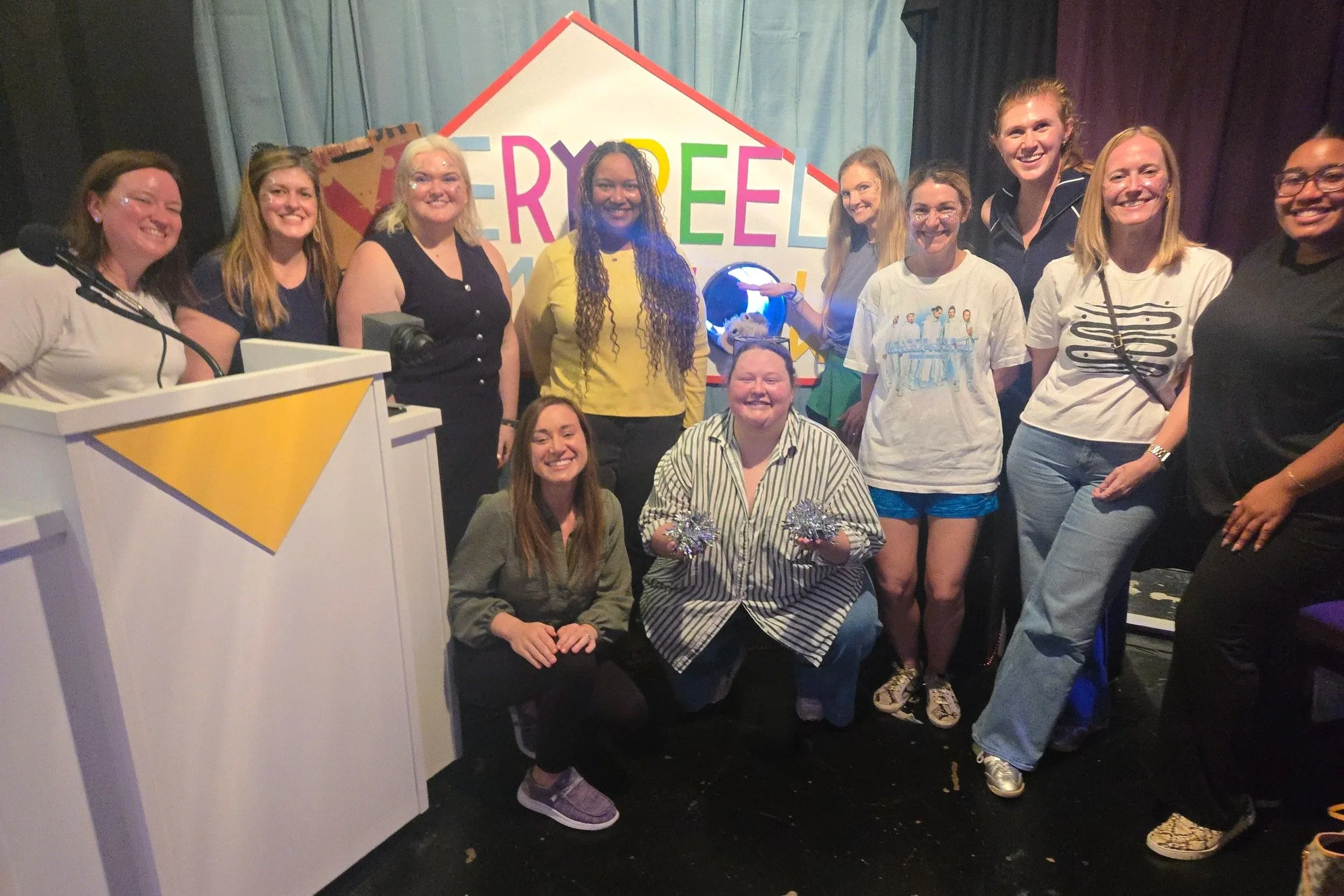 Group of smiling people, mostly young women, standing together in front of the Very Reel Game Show sign with a puppet rat. The atmosphere appears lively and celebratory.