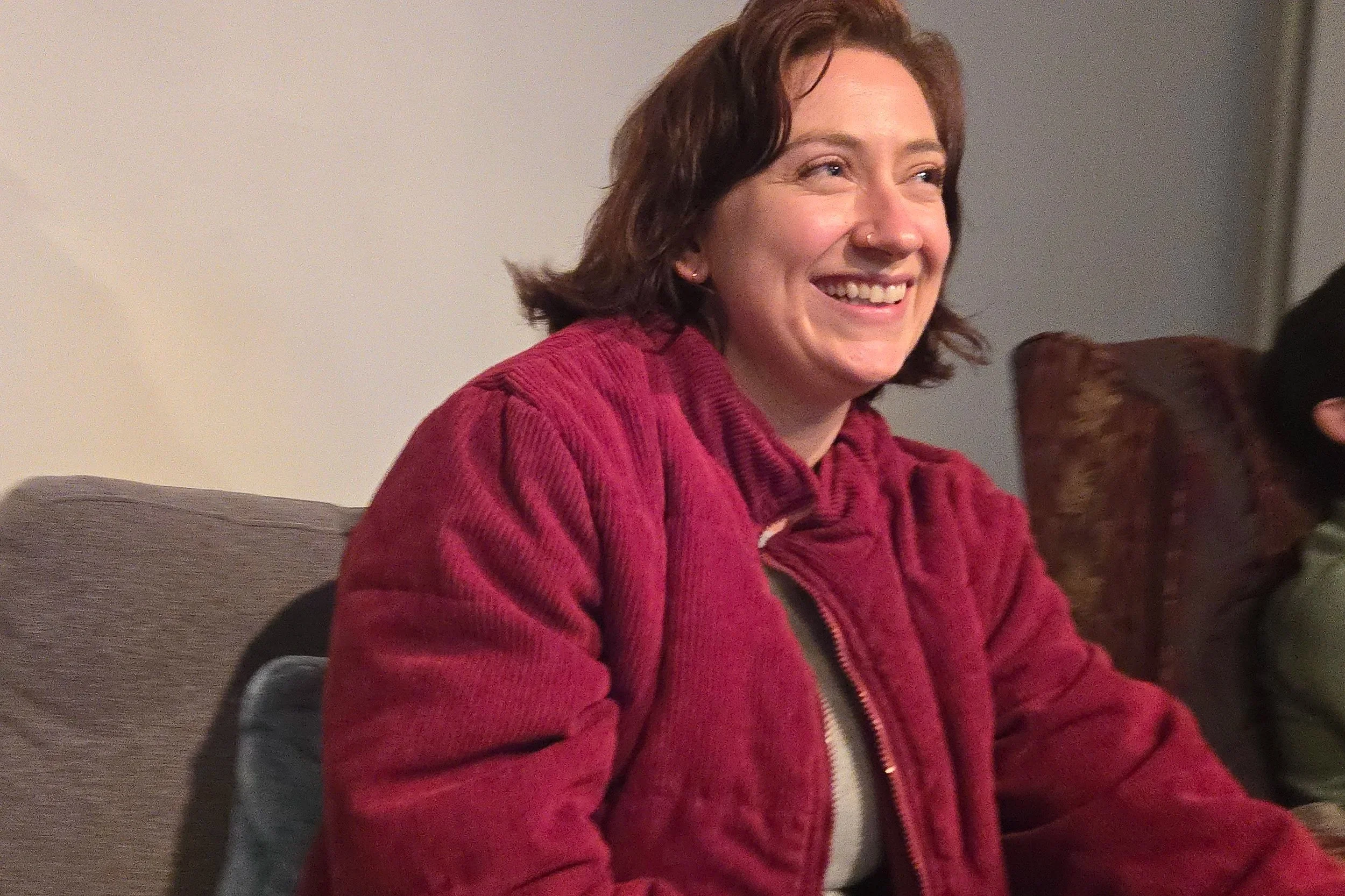 A woman with short brown hair, wearing a red jacket, smiling and sitting in a room.